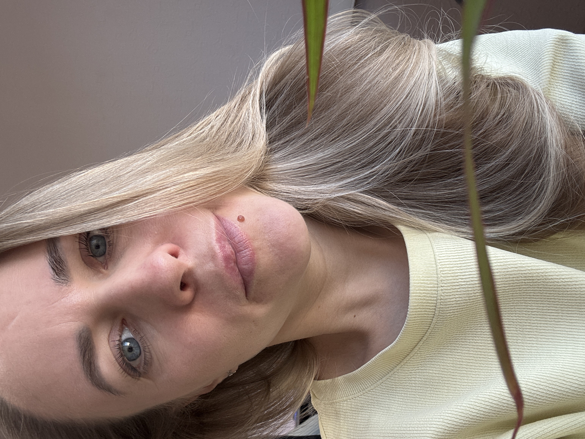 A close-up, angled portrait of a fair-skinned woman with blonde hair and blue eyes. She is wearing a light yellow ribbed sweater. A plant stem with a reddish-green leaf partially obscures the view. The background appears to be a light gray wall. There is no identifiable location in Chaikovsky, Russia, in the image.