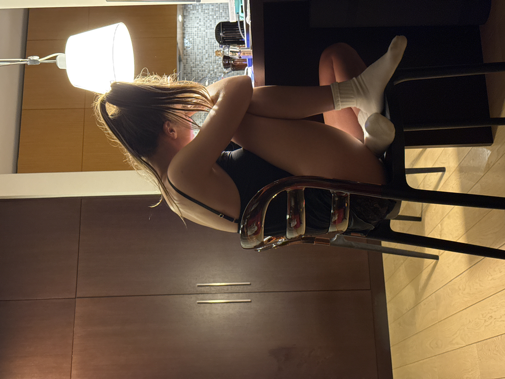 A young woman with brown hair pulled back in a ponytail is sitting in a chair. She is wearing white socks and a dark-colored bodysuit. Her legs are pulled up to her chest, and her arms are wrapped around them. The chair has a chrome back and black legs. Behind her is a light-colored wooden wall, and a lamp is positioned above her head. In the background, a glimpse of a kitchen counter can be seen. The floor is made of light-colored wooden planks. The location appears to be an apartment interior, possibly in Warsaw, Poland, although there is no specific evidence in the image to confirm the city.