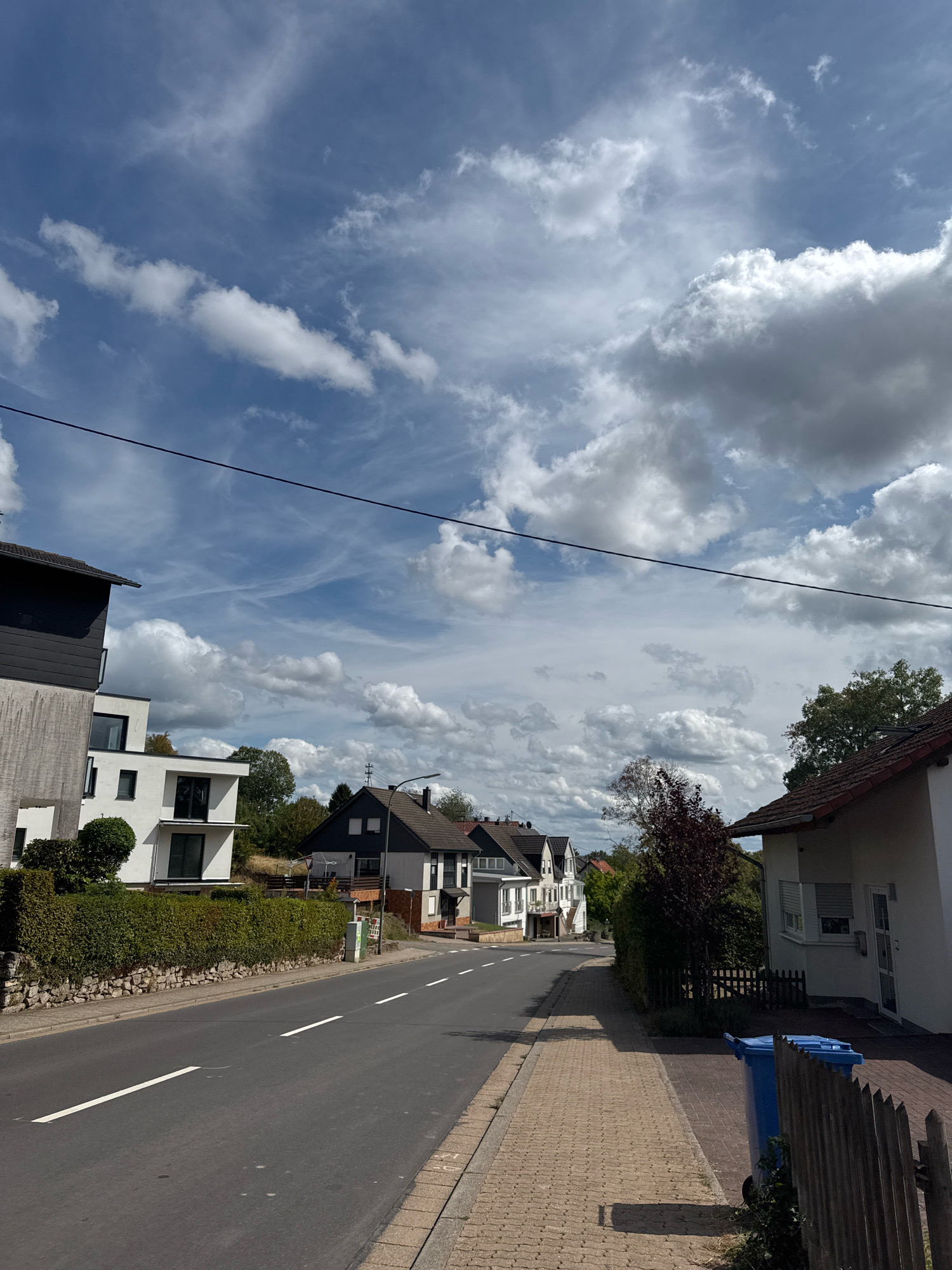 The image depicts a residential street in Überherrn, Germany, under a partly cloudy sky. The street is paved with asphalt and has white lane markings. To the left of the street, there is a stone wall topped with a neatly trimmed green hedge, and behind it, several modern-style houses with white facades and dark roofs. One of these houses features a multi-story design with large windows. Further down the street, more houses with traditional dark roofs are visible, interspersed with trees. To the right of the street, there is a paved sidewalk with a textured surface. Adjacent to the sidewalk, a white house with a red-tiled roof is visible, featuring white shutters on its windows. A wooden fence runs along the property line, and two blue trash bins are placed near the sidewalk. The environment appears calm and suburban, with no people or vehicles present in the scene.
