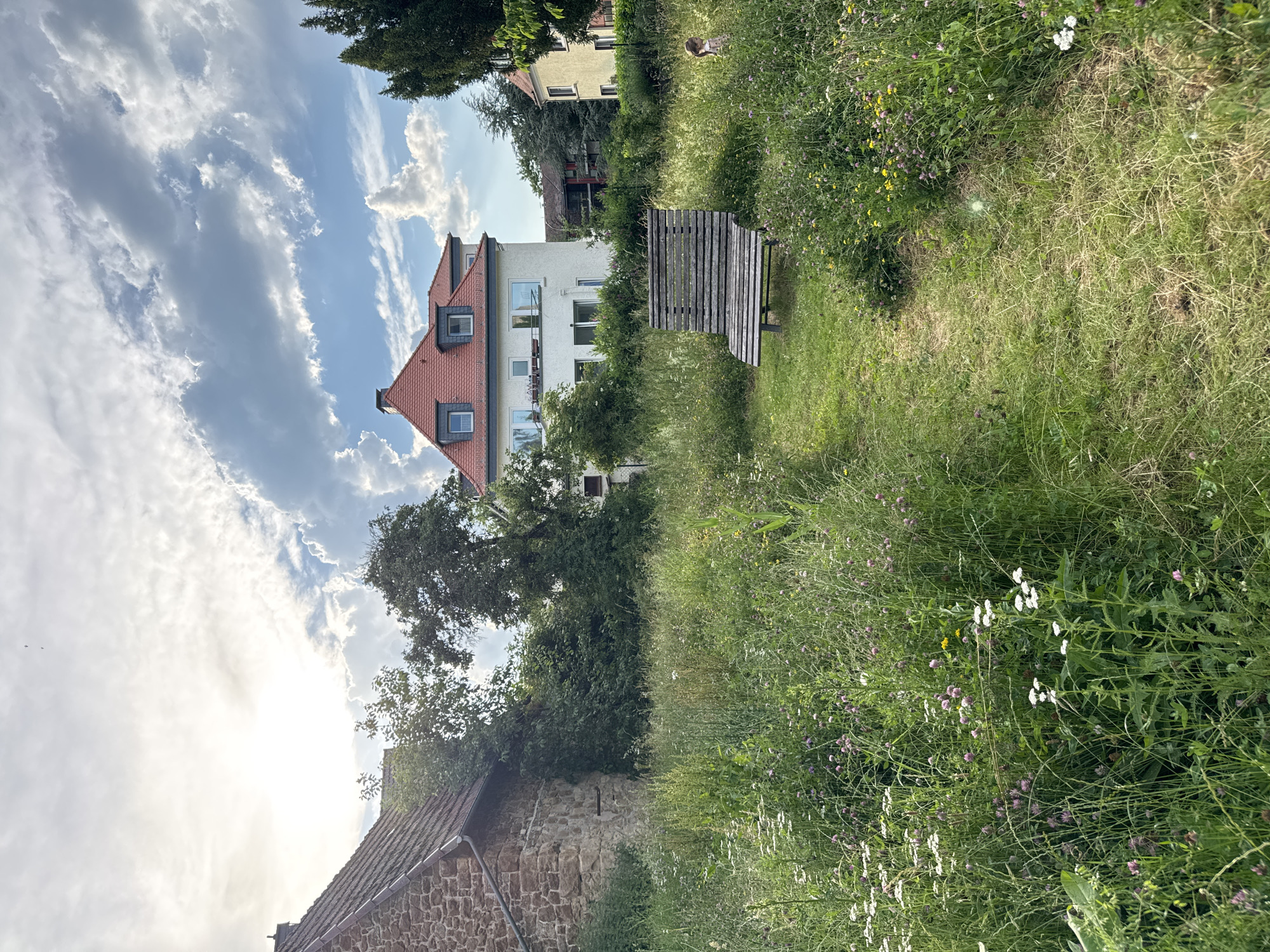 A rustic wooden bench sits in a meadow of tall grass and wildflowers. The bench is worn and weathered, with a slatted seat and back. It is positioned on a dirt path that winds through the meadow. In the background, a large, white building with a red-tiled roof is visible. The building has several windows and balconies. To the left of the bench, a stone building with a similar red-tiled roof can be seen. The sky is partly cloudy, with the sun peeking through the clouds. A small child with blonde hair, wearing a pink and white dress, is visible to the right of the bench, partially obscured by the tall grass. The overall scene evokes a sense of nature reclaiming an urban or semi-urban space.