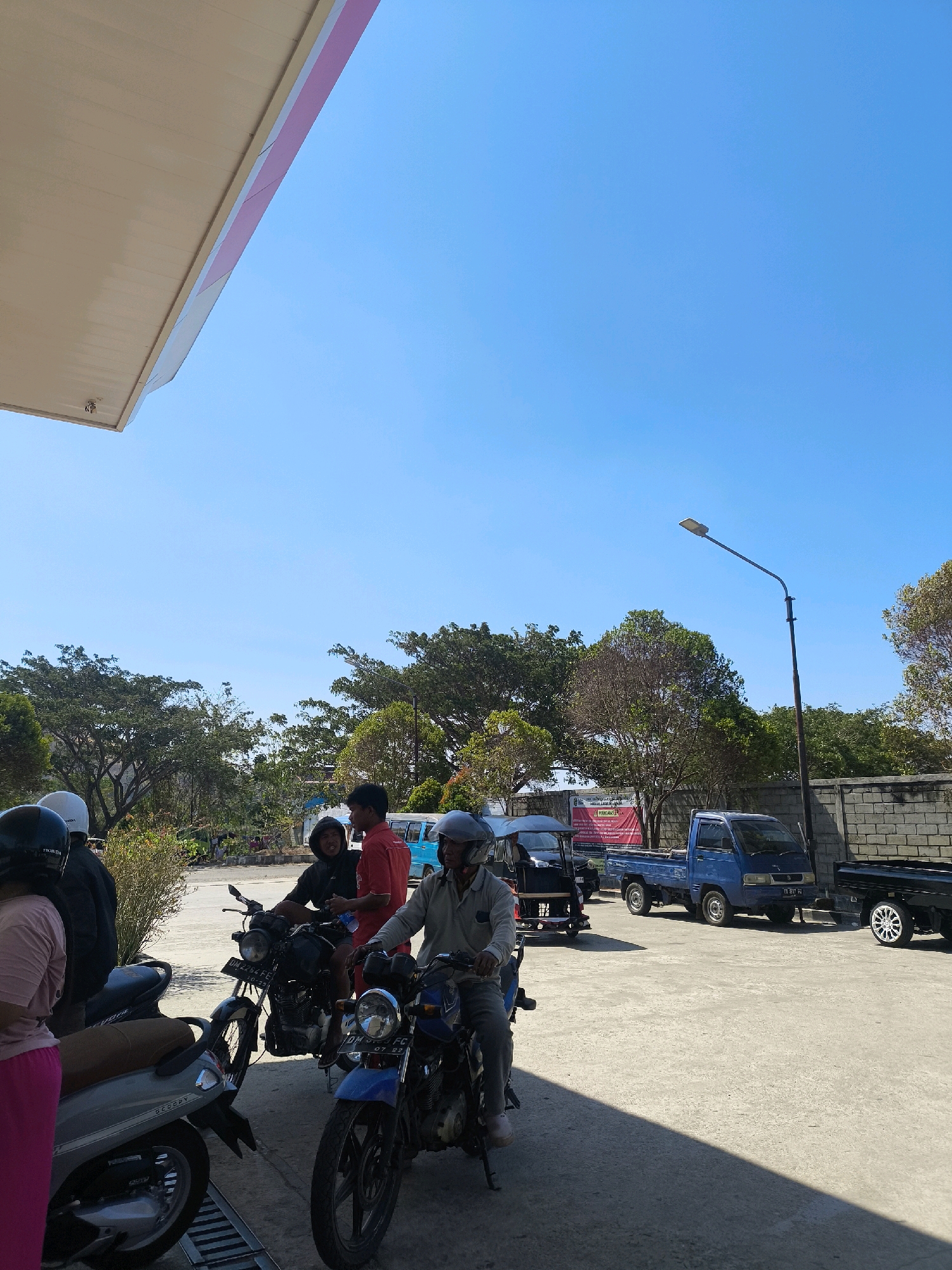 The image depicts a bright, sunny day at a gas station in Kwandang, Indonesia. Several individuals are present, some on motorcycles. In the foreground, two motorcycles are parked. One is a blue motorcycle with a rider wearing a gray long-sleeved shirt and a helmet. Another is a black motorcycle with a rider wearing a black hooded jacket and a helmet, holding a bottle of water. To the left, a person in a pink outfit and a helmet is partially visible. In the background, a blue pickup truck is parked, and further back, a black truck with distinctive silver rims is seen. Trees and a brick wall line the perimeter of the gas station. The sky is a clear, vibrant blue. The overall scene conveys a casual, everyday moment at a local establishment.