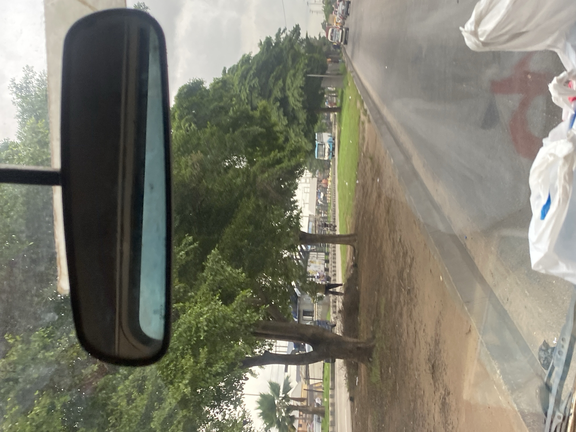 The image shows a view from inside a vehicle looking through the windshield. A rearview mirror is visible at the top center of the frame, reflecting trees and the sky. Outside the vehicle, a street scene in Lagos, Nigeria unfolds.
The street is lined with trees on the left, with a sidewalk and a median. Beyond the median, there is a road with several vehicles, including a blue and white bus, a white mini-bus, and cars. People can be seen on the side of the road and near the buildings in the background. The buildings appear to be commercial or residential structures.
The ground on the left side of the street is unpaved and covered with dirt and some scattered debris. The road itself is paved and shows signs of use. The sky is overcast, suggesting a cloudy day.
In the foreground, inside the vehicle, the lower part of the windshield is visible, with some smudges and reflections. There are white plastic bags in the lower right corner of the frame, suggesting personal belongings or cargo. A red painted symbol, possibly a traffic marking or graffiti, is visible on the windshield.
The overall impression is a bustling street scene in a developing urban environment.