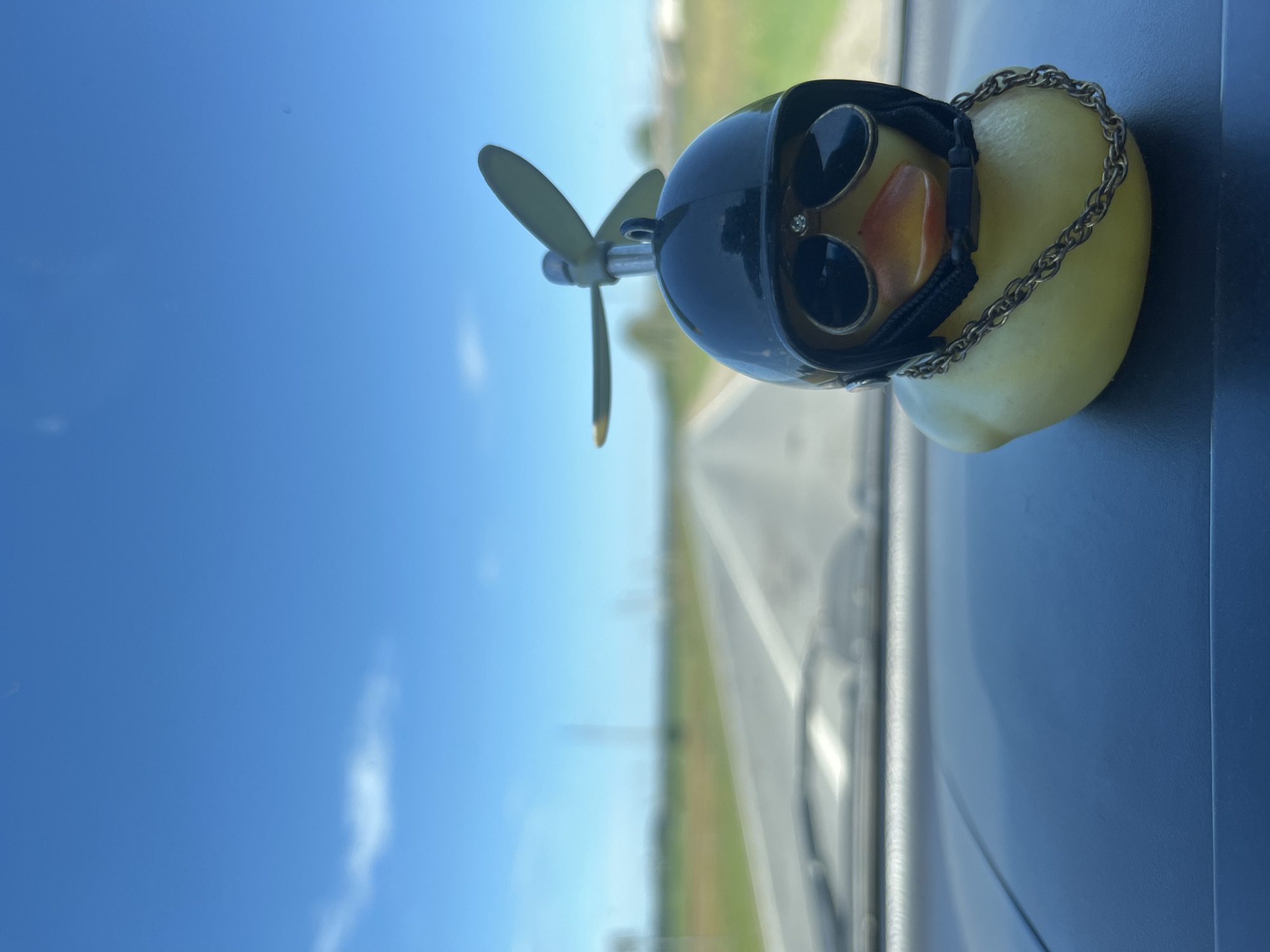A yellow rubber duck toy adorned with a black helmet, sunglasses, and a gold chain is positioned on a dark dashboard in the foreground. The duck has a propeller attached to its helmet. The background shows a blurry asphalt road stretching into the distance, flanked by green fields and a clear blue sky with scattered clouds. The scene appears to be taken from inside a vehicle, suggesting a road trip. The location in Kukshumy, Russia, is not discernible from the visual information provided in the image.