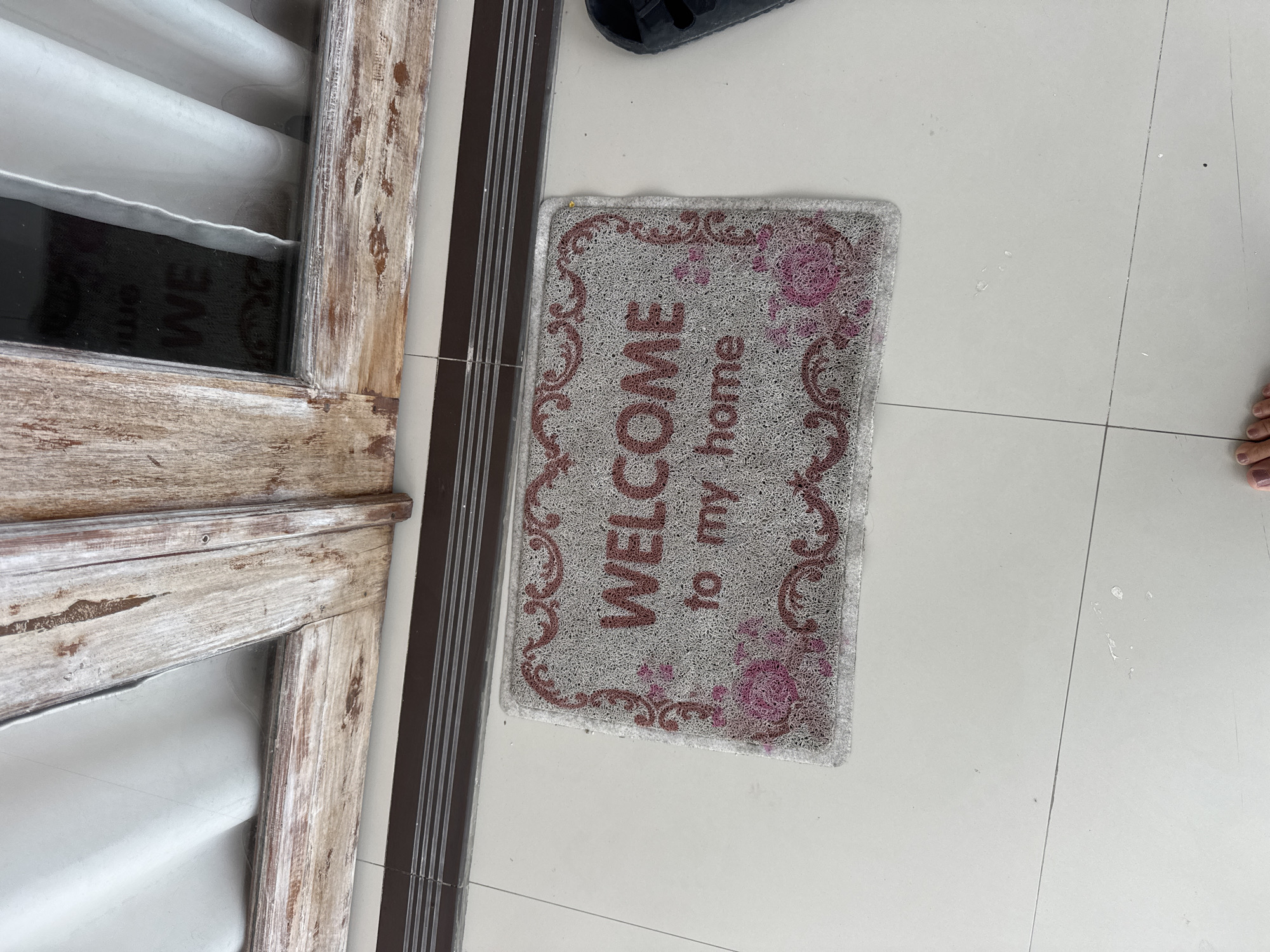 A welcome mat reading "WELCOME to my home" is situated on a light-colored tiled floor in front of a weathered wooden doorframe. The doorframe appears to be part of an entrance to a building, with a white curtain visible through the glass panels of the door. A black sandal is placed to the right of the mat, partially visible in the frame. The scene is captured from a slightly elevated perspective, showing a small portion of a person's feet at the bottom of the image. This appears to be the entrance to a residence or a commercial establishment in Nusa Penida, Indonesia.