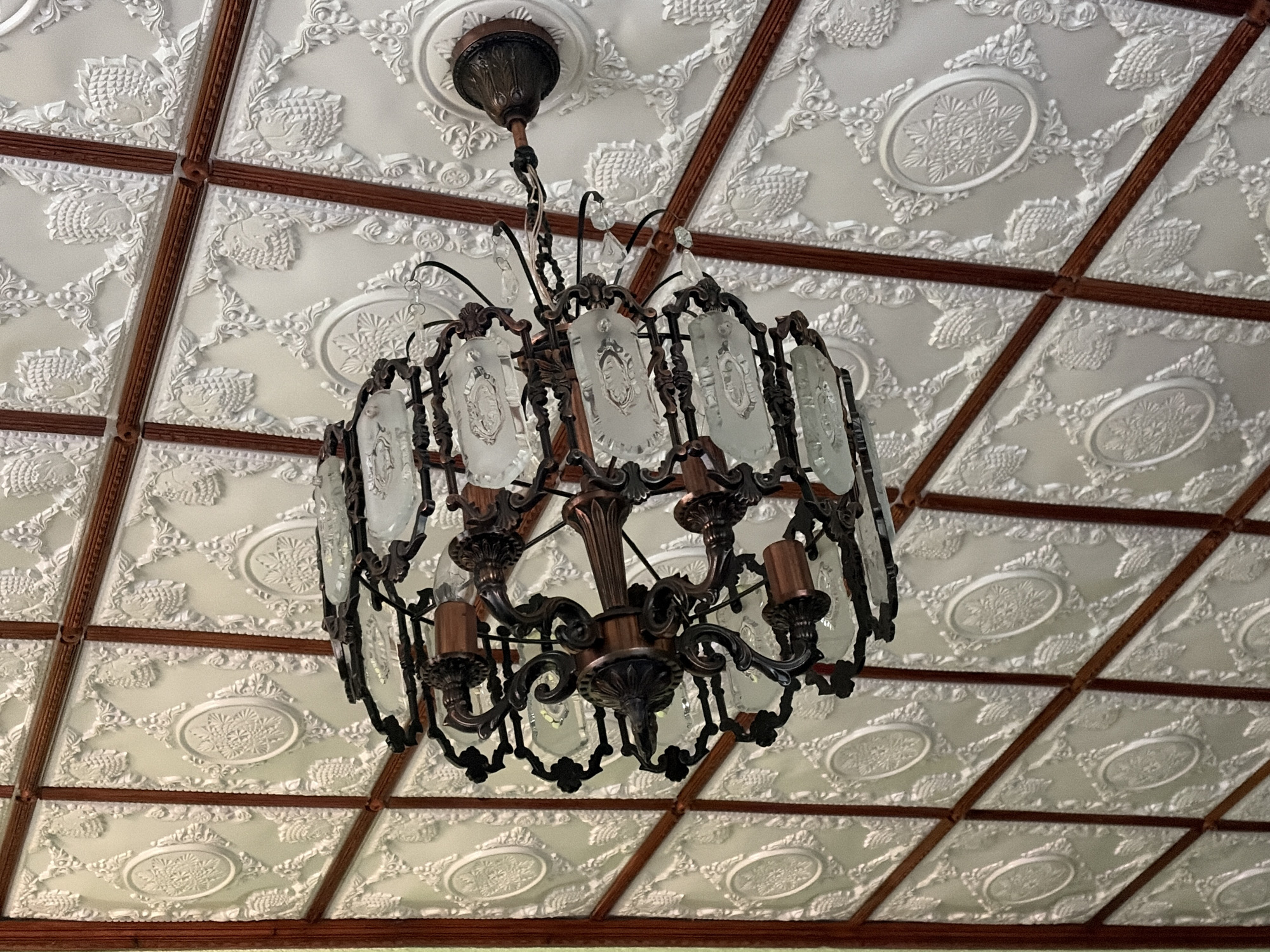 The image shows a close-up, low-angle shot of an ornate chandelier hanging from a decorative embossed ceiling in what appears to be a well-lit interior. The ceiling is white with intricate raised patterns, featuring floral motifs and oval medallions. Dark brown wooden beams divide the ceiling into a grid. The chandelier itself is made of dark metal, with multiple arms holding glass shades and some decorative crystal drops. The glass shades are rectangular with frosted or etched designs. The overall impression is one of vintage or classic interior design. No specific location in Hanniv, Ukraine is identifiable from the image.