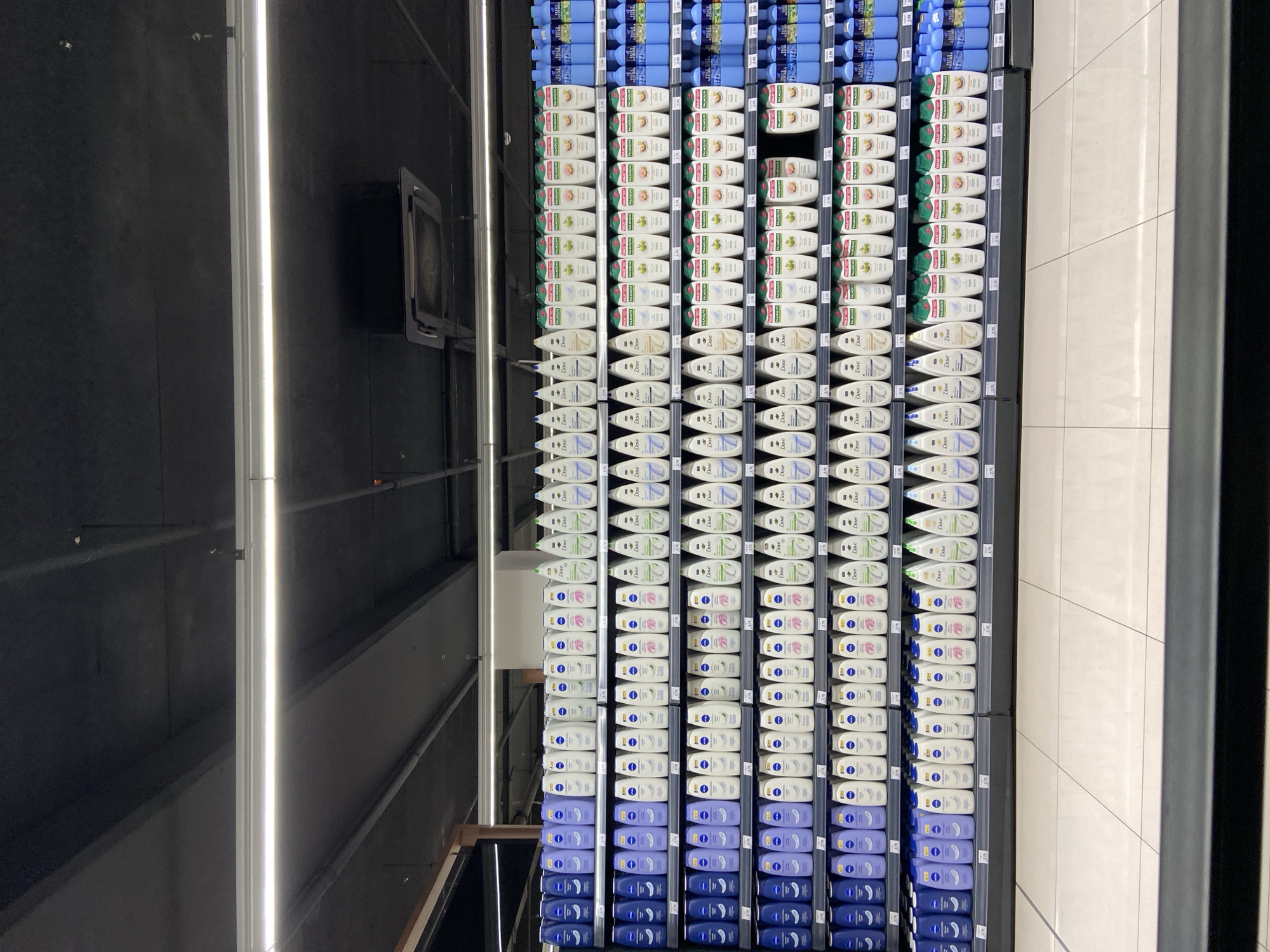 The image depicts a well-stocked supermarket shelf, probably in Budva, Montenegro, with rows of body wash bottles arranged by brand and scent. The shelves are filled with similar products, neatly aligned, with white and blue being the dominant colors, indicating different product lines. The overhead lighting suggests an indoor setting, possibly a retail store in Budva. The camera angle is likely from the perspective of a shopper or someone stocking the shelves.