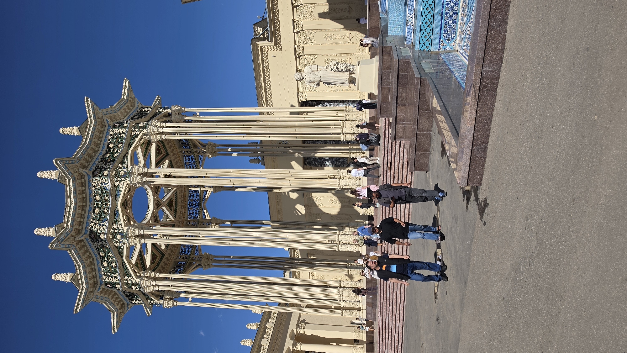 The image depicts the Uzbekistan Pavilion at VDNKh in Moscow, Russia. A large, ornate pavilion with a multi-tiered roof and numerous slender columns stands against a clear blue sky. The pavilion's architecture is intricate, featuring decorative latticework and detailed moldings. In the foreground, three men are walking on a paved area. The man on the left wears a black vest over a blue shirt and jeans. The man in the center wears a black t-shirt and jeans. The man on the right wears a grey t-shirt and dark pants. Behind them, a set of tiered red steps leads up to the pavilion. Further back, various people are visible walking and standing, some near the steps and others in front of the pavilion's facade. To the right of the pavilion, a white statue of a woman holding a bouquet of flowers stands on a pedestal. In the very right of the image, a section of a fountain with blue mosaic tiles is visible. The overall scene is well-lit, suggesting it was taken on a sunny day.