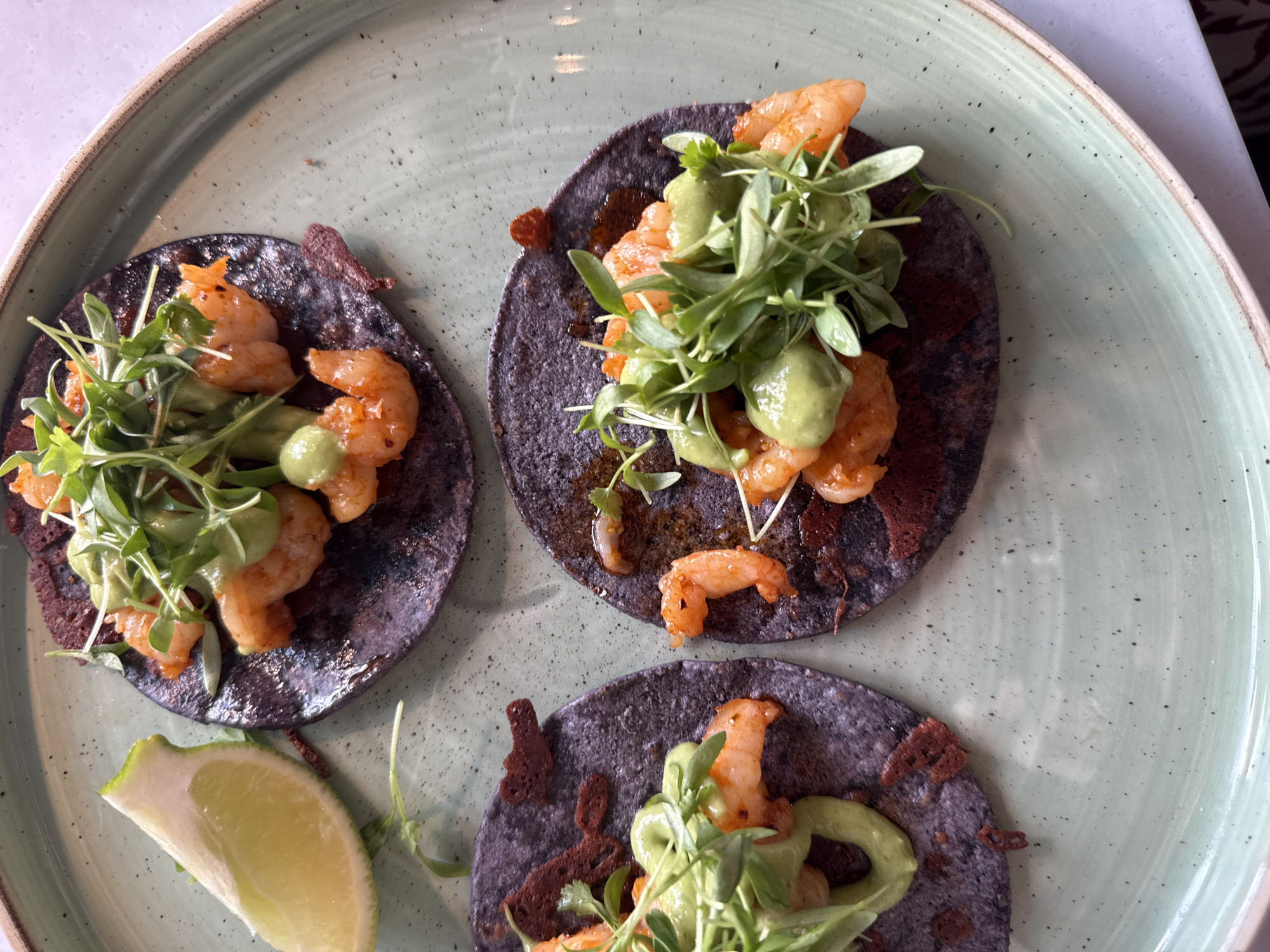 The image shows a plate of three shrimp tacos, arranged in a roughly triangular pattern. The tacos are made with what appear to be blue corn tortillas. Each taco contains cooked shrimp, a green sauce (possibly guacamole or a similar avocado-based sauce), and microgreens. There are some pieces of dark crispy food on the tortillas, likely some sort of garnish. A wedge of lime is present on the plate next to one of the tacos. The plate itself is a light green color with a speckled pattern and a slightly raised rim. The background is slightly out of focus, suggesting a restaurant setting. Given the location of Solana Beach, United States of America, the image may have been taken inside a restaurant located in that city.