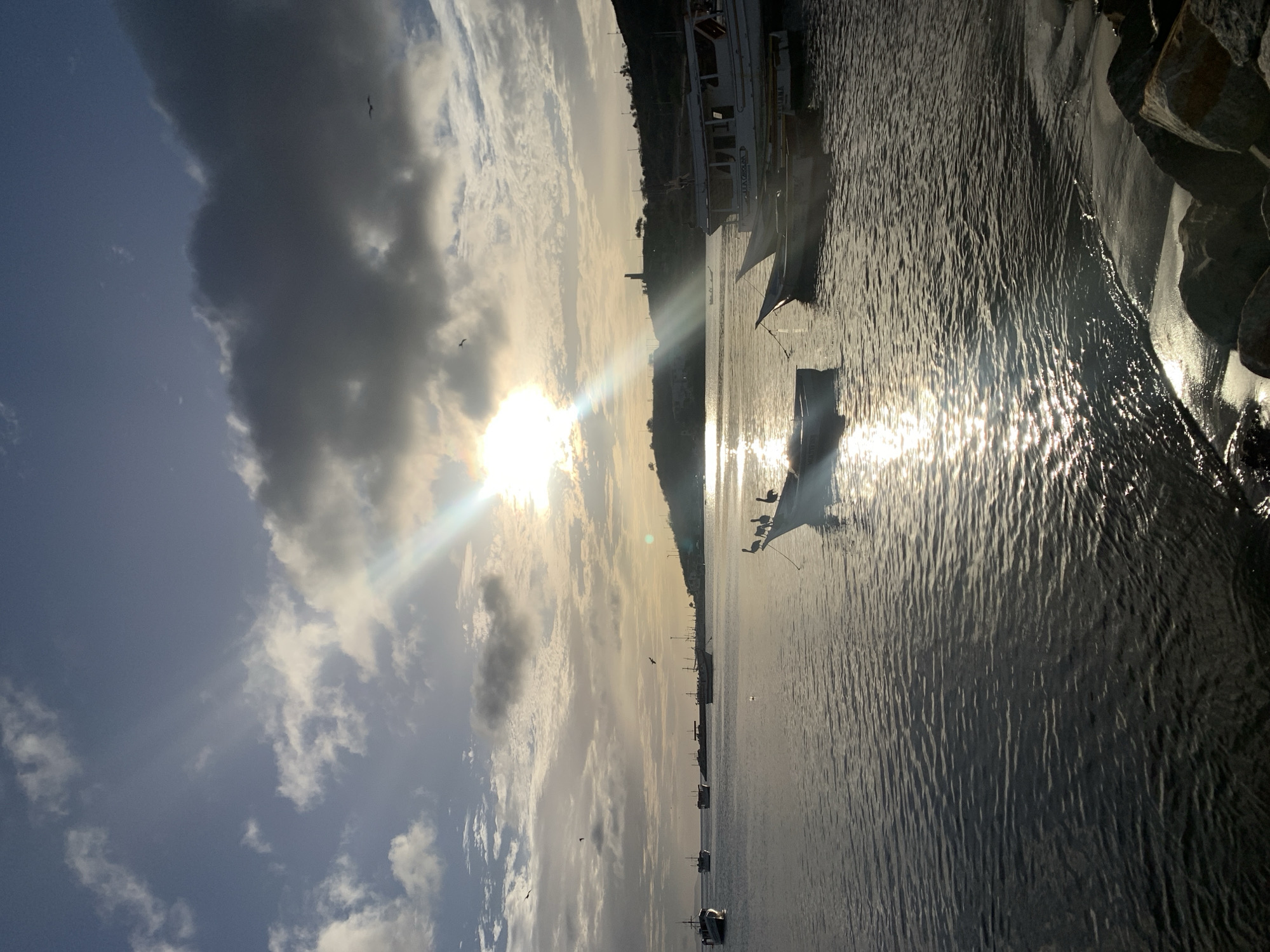 The image depicts a serene coastal scene at Juan Griego, Venezuela, during what appears to be either sunrise or sunset. The sky is a mix of blue with scattered clouds, some of which are dark and dramatic, while others are wispy and illuminated by the sun. Sunbeams dramatically cut through the clouds and reflect off the water, creating a shimmering effect.
Several boats are anchored in the calm bay. In the foreground, a white boat is prominently featured with three pelicans perched on its bow. Further out in the bay, other boats of varying sizes are visible, some appearing to be fishing vessels. In the distance, the coastline is lined with hills and some buildings, including a tower-like structure on one of the higher points.
The water is relatively still, with gentle ripples on the surface. The immediate foreground shows a rocky shoreline with some sand. The overall atmosphere is peaceful and tranquil, capturing the natural beauty of the location. There are no people clearly visible in the image. The lighting suggests a time of day with low sun angle, enhancing the atmospheric qualities of the scene.