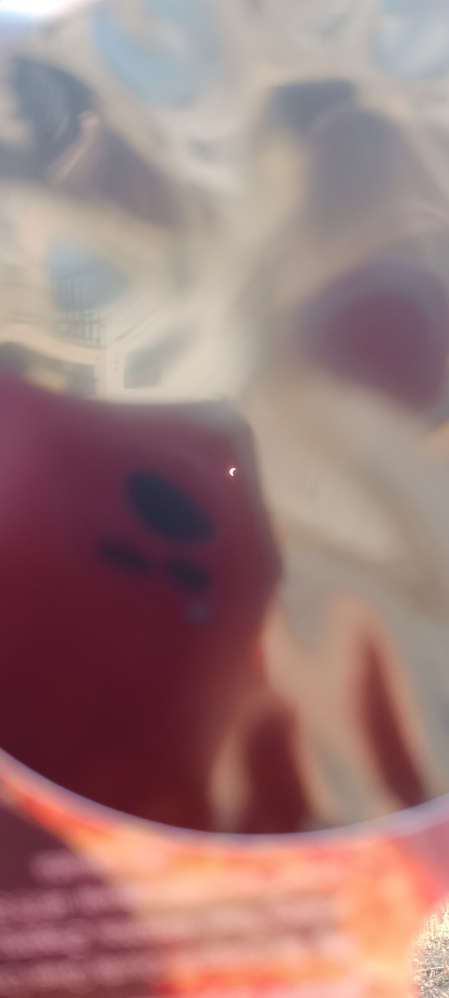 The image shows a close-up, slightly blurry view of a red object, possibly a smartphone, reflecting a bright light source, potentially the sun during an eclipse. The reflection shows a crescent shape, indicating that the sun is partially obscured. The background is a mottled pattern of light and dark colors, suggesting a textured or layered surface. The scene does not contain any recognizable landmarks or elements that would indicate its specific location within Adai, Spain.