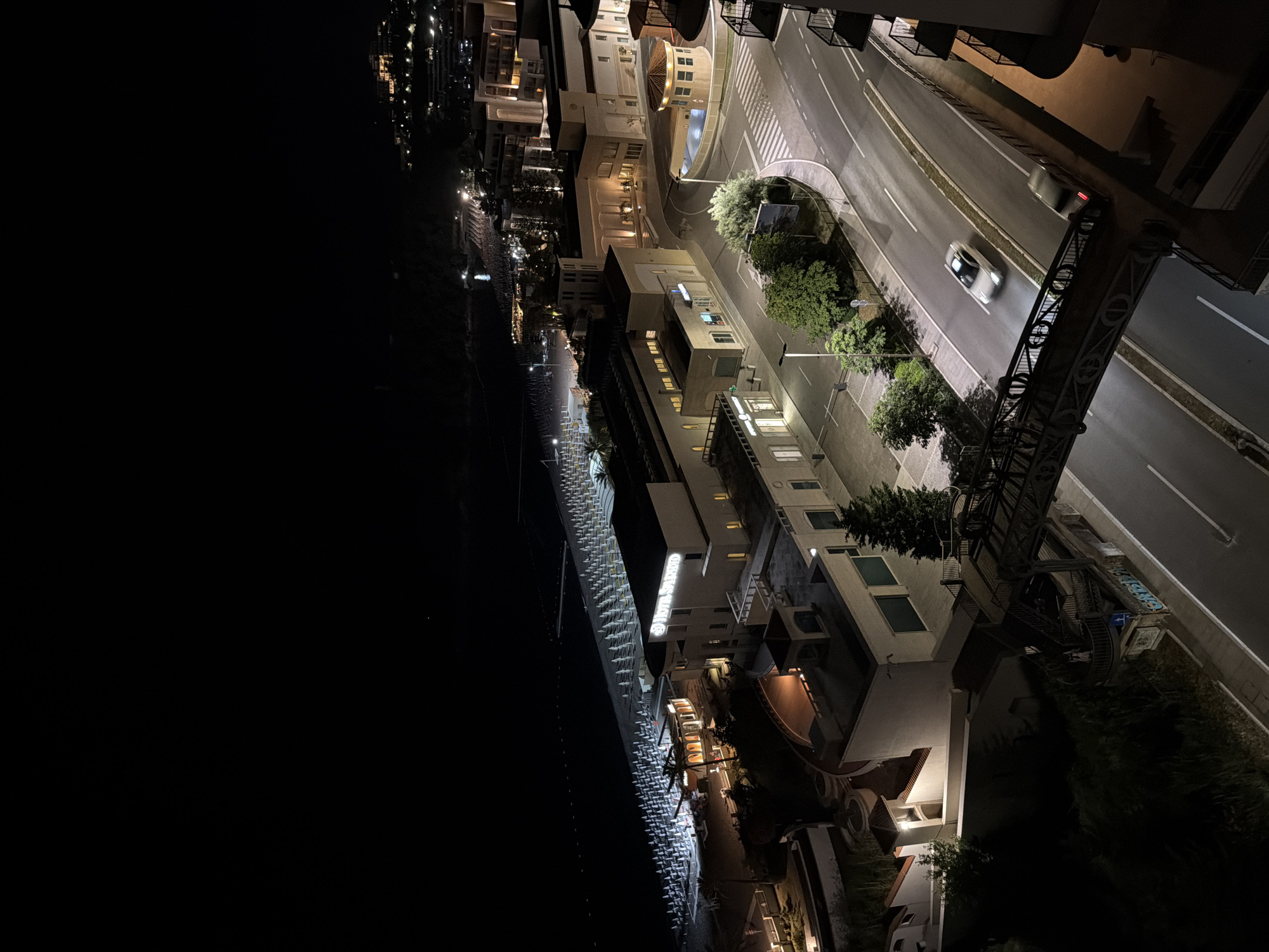 This is a high-angle, overhead night photograph of a coastal urban area in Boreti, Montenegro, illuminated by artificial light sources. The scene captures a multi-lane asphalt road in the foreground, traversed by a dark metal pedestrian bridge with ornate railings. Two vehicles are present on the road: a light-colored car is in motion, exhibiting significant motion blur, while a darker vehicle is stationary or moving slowly in the opposite direction.
Adjacent to the road are various commercial and residential buildings with light-colored facades. On the right side of the road, a complex of buildings includes structures with illuminated windows and a prominent, illuminated sign displaying "HOTEL SPLENDID" on one of its larger edifices. A roundabout is visible, featuring a small, circular, tiled-roof structure in its center.
To the left of the main road and buildings, a wide beach extends along the coastline. The beach is densely arranged with numerous rows of white sun umbrellas and corresponding sunbeds, all brightly illuminated. The adjacent sea is dark, reflecting some of the shore lights. Further along the coastline, more illuminated buildings ascend a hillside, indicative of continuous development. The sky is uniformly dark, and no individuals are discernibly visible within the frame. Streetlights, building lights, and beach lights are the primary sources of illumination for the entire scene.