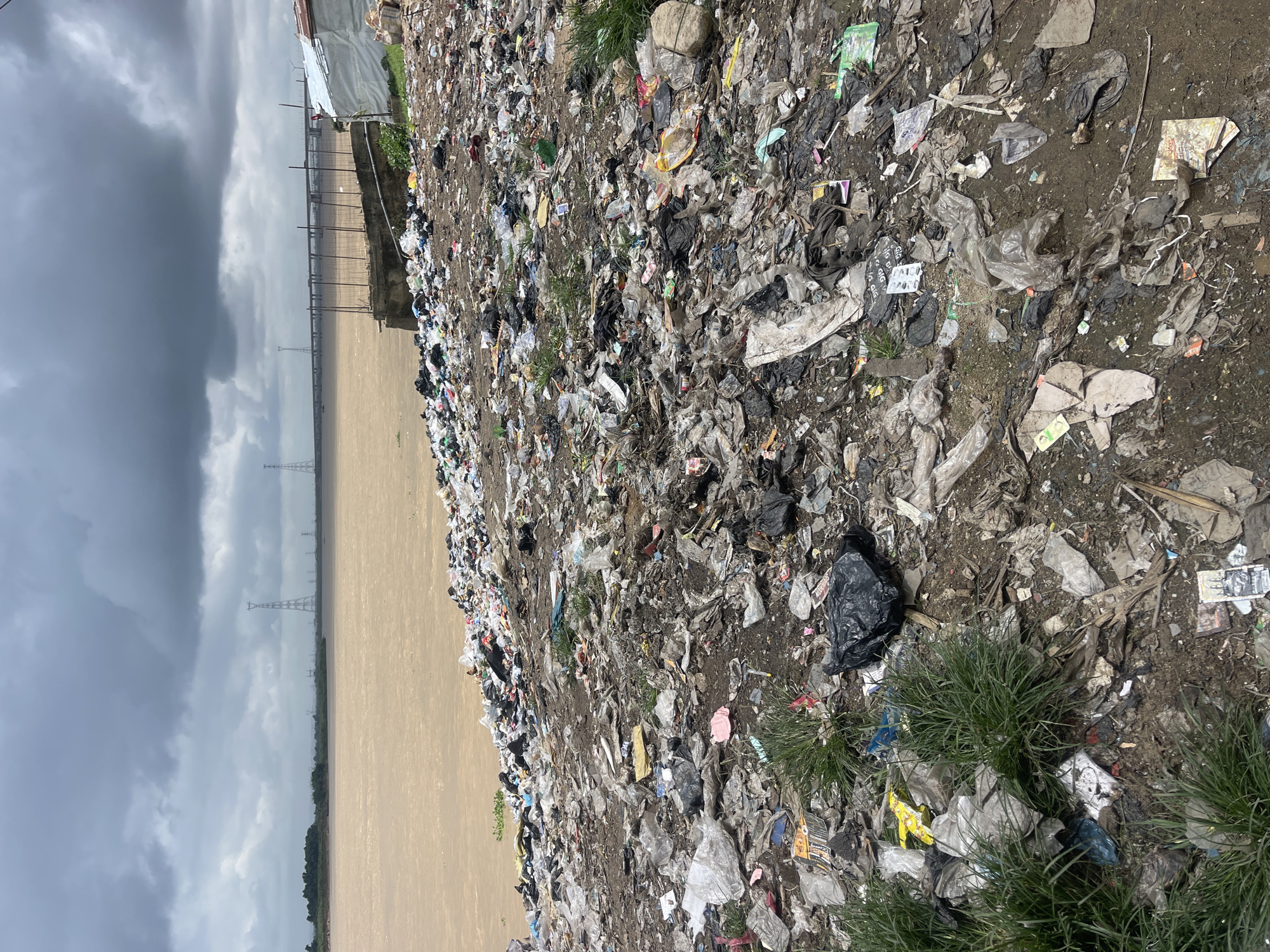 The image shows a heavily polluted riverbank in Atani, Nigeria. The foreground is covered in a thick layer of trash, including plastic bags, bottles, paper, and other debris. Some patches of green grass struggle to grow through the garbage. In the background, a wide, muddy river flows under a cloudy sky. Power pylons and a bridge are visible in the distance, hinting at the presence of a larger urban area. A makeshift shelter made of white sheeting is visible on the right side of the image, suggesting a human presence in this otherwise desolate environment. The overall scene depicts severe environmental degradation and a lack of waste management infrastructure.