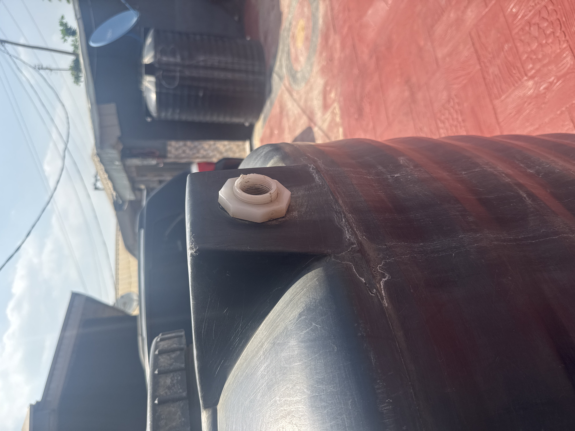 This is a close-up, angled shot of the exterior of a large, dark-colored plastic water tank. The tank has a ribbed texture visible on its surface, with a significant amount of surface scratching and some lighter-colored material flaking off in places, suggesting wear and tear. Prominently featured on the upper portion of the tank is a hexagonal, white plastic fitting with internal threading, likely an outlet or inlet connection. The fitting appears to have some dirt or residue on it.
The background is out of focus but reveals elements of the surrounding environment. To the right, a red, textured surface is visible, possibly a wall or floor, with a circular pattern embossed or painted on it. To the left, a bright sky with some clouds suggests daylight. A faint reflection of trees and a power line is visible on a shiny surface in the upper left quadrant, potentially another tank or a reflective building. In the distance, hints of buildings and structures can be discerned, along with what appears to be other stacked water tanks.
The scene appears to be outdoors, likely in a residential or commercial area where water storage is common. The lighting suggests it is daytime, possibly late morning or afternoon given the brightness and shadows. There are no people visible in the frame, and the focus is solely on the water tank and its immediate surroundings. The overall impression is one of utilitarian infrastructure, possibly in a region with water supply challenges. There is no visible text in the image.