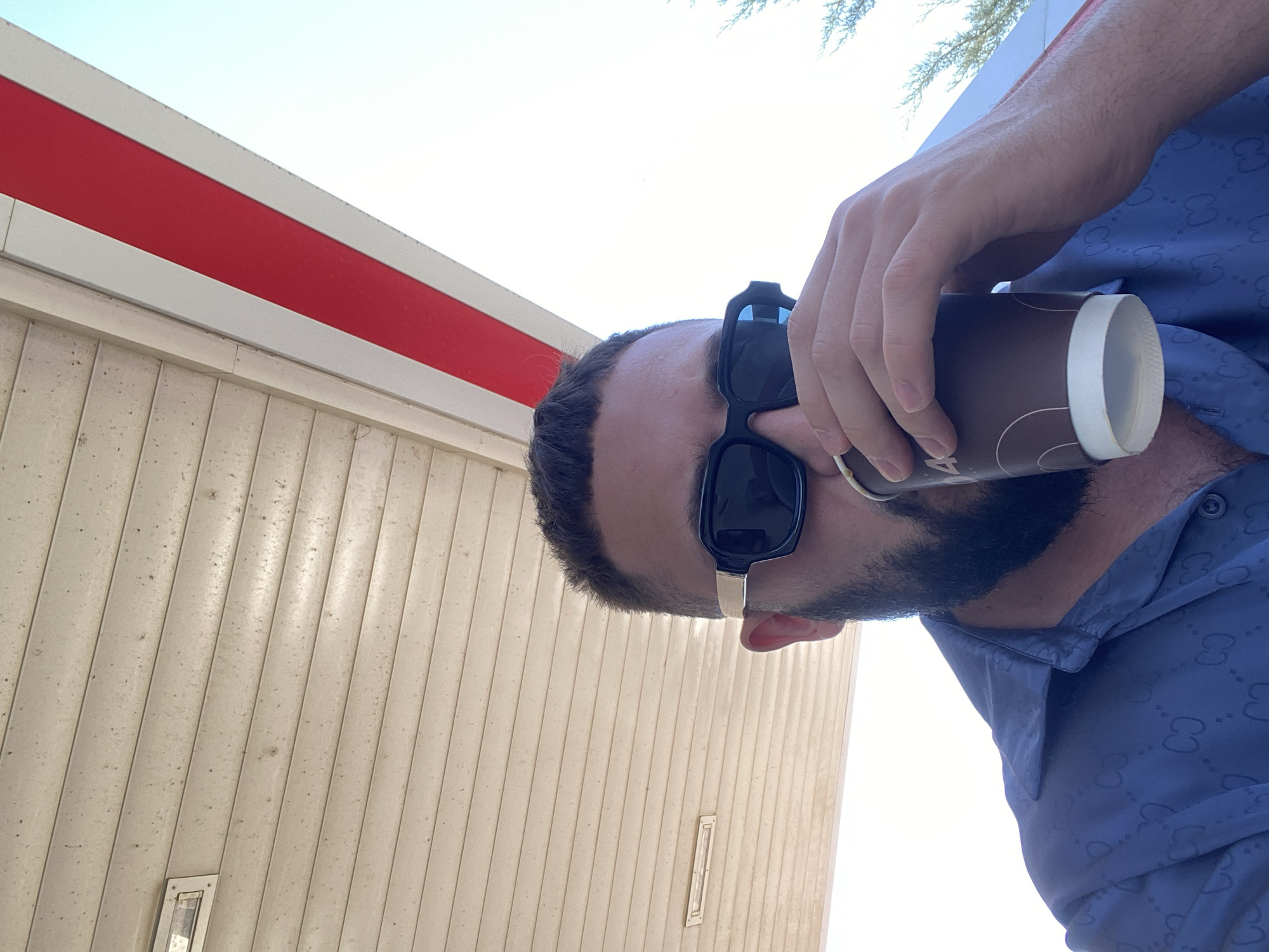 In the city of Korkmaskala, Russia, a man with a beard and sunglasses is seen drinking from a coffee cup. He is wearing a blue button-down shirt with a repeating pattern. The man is positioned under an awning, with a light-colored ceiling above and a bright sky visible. The awning has red and white striped accents.