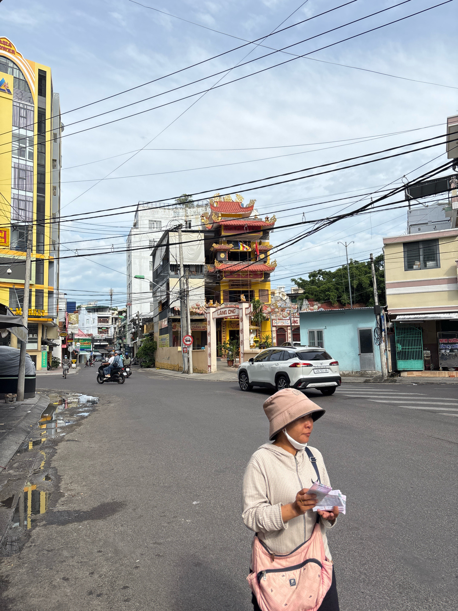 The image shows a street scene in Nha Trang, Vietnam. In the foreground, a woman wearing a light-colored hooded sweatshirt, a bucket hat, and a face mask is standing on the street, holding a wad of cash in her hands. She has a pink cross-body bag slung over her shoulder. Behind her, a white SUV is driving across a pedestrian crossing. To the left, a bright yellow building with arched windows and a sign that reads "ASIA TOWER" is visible. Further down the street, a temple-like building with ornate red and gold roofs stands prominently. The street is paved and shows signs of recent rain, with puddles reflecting the surrounding buildings. Several utility poles with a tangle of wires are present, characteristic of urban Vietnamese streetscapes. Motorcycles and other vehicles can be seen in the background, indicating a busy urban environment. The sky is overcast with scattered clouds.