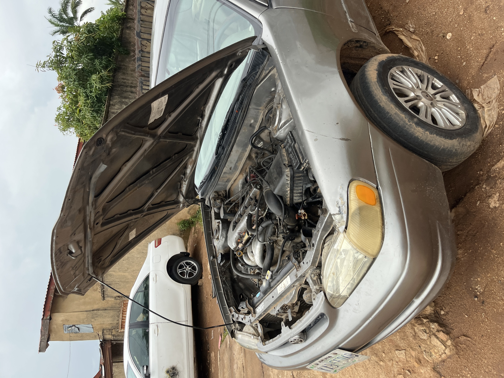 In Bukuru, Nigeria, a silver sedan with its hood open is parked on a dirt surface. The engine compartment is exposed, revealing various mechanical components. Behind the silver car, a white sedan is visible, also with its hood down. The background features a building with peeling paint and a concrete fence with vegetation growing over it. The sky is overcast. The license plate of the silver car is visible and reads "XAB 504R".