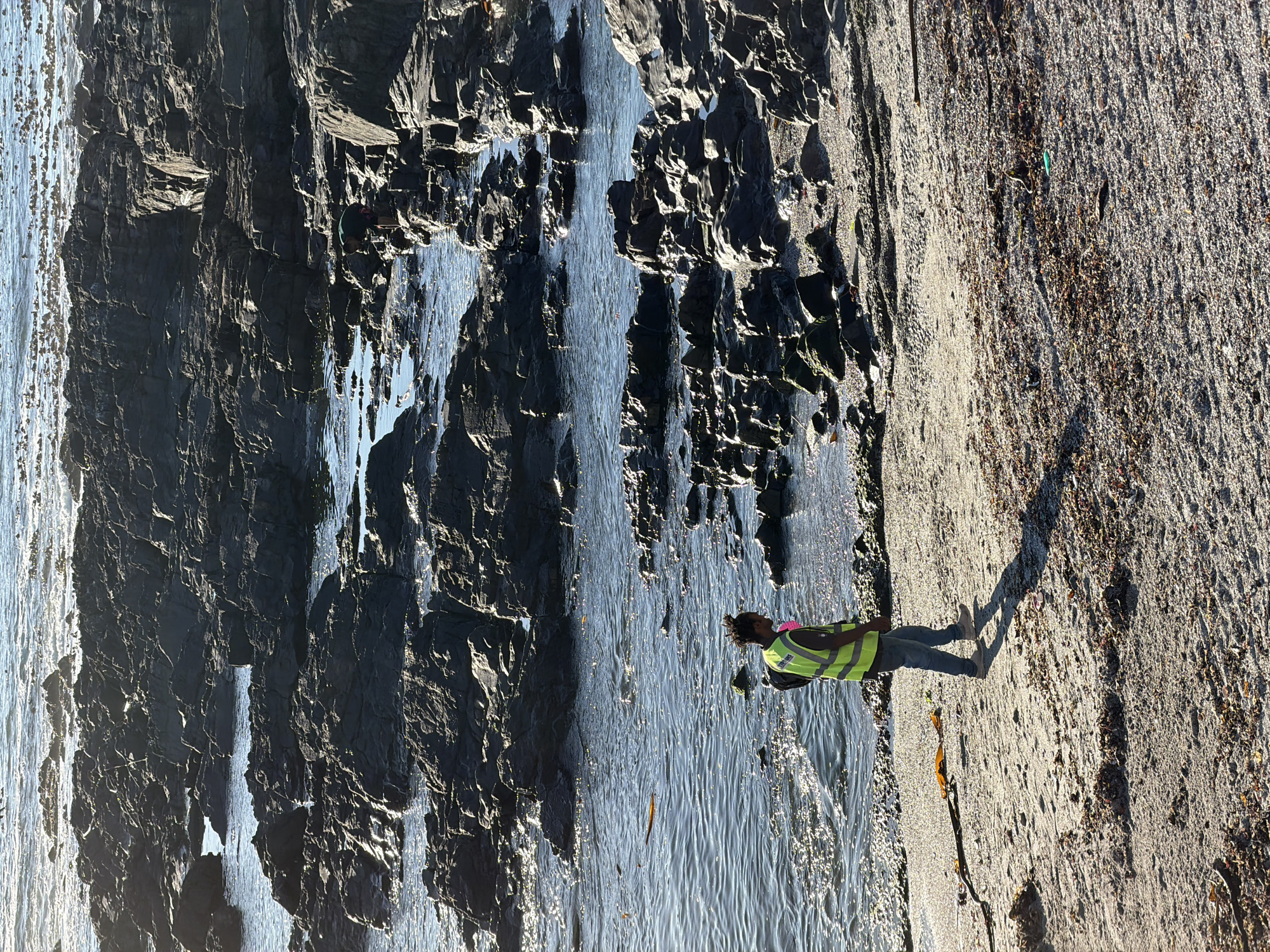 A vertical-format, medium-shot photograph depicts a woman in a neon yellow safety vest standing on a sandy beach near a rocky cliff face. The woman is wearing jeans and boots, and appears to be looking down at the beach. The cliff face is dark gray and appears to be wet. The ocean is visible in the background. The scene is bathed in bright sunlight, casting a long shadow from the woman onto the sandy beach. The location is likely near the shoreline of Cape Town, South Africa, given the combination of rocky cliffs, sandy beach, and ocean.
