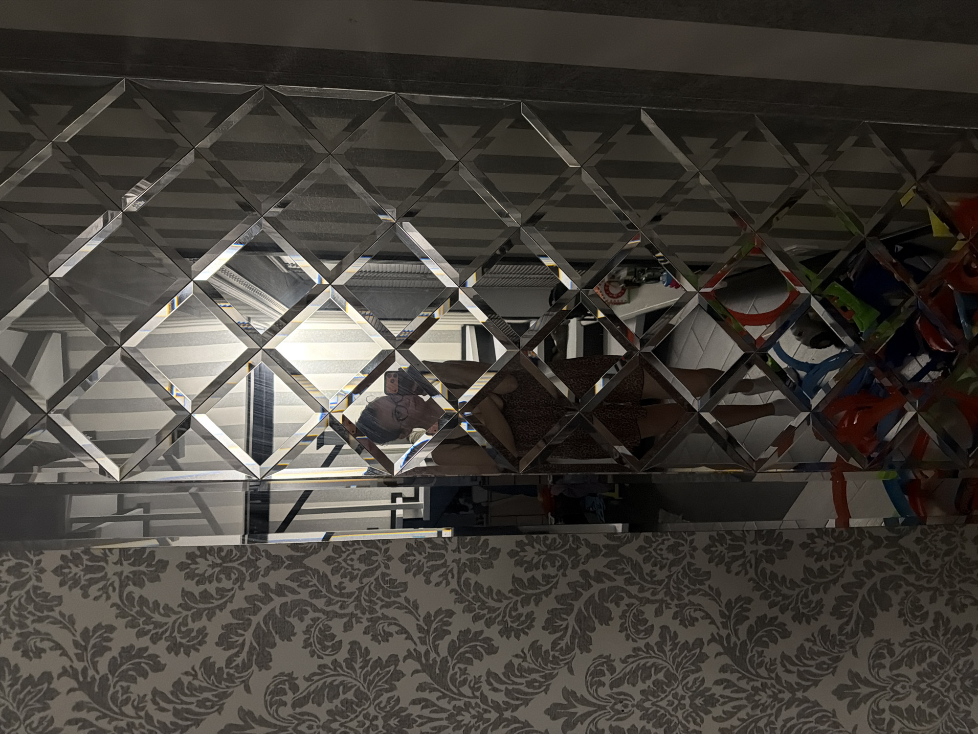 The image shows a woman taking a selfie in a mirrored wall. The wall is composed of diamond-shaped mirrored panels. To the left of the mirrored wall is a wall covered with damask-patterned wallpaper. In the reflection, the woman is wearing glasses and a dress. Her legs and feet are visible. Behind her, the reflection shows a room with striped wallpaper and some objects on the floor, including what appears to be toys. The scene suggests an indoor residential setting, possibly a hallway or living area. There is no information in the image to indicate the location is in Bila Tserkva, Ukraine.