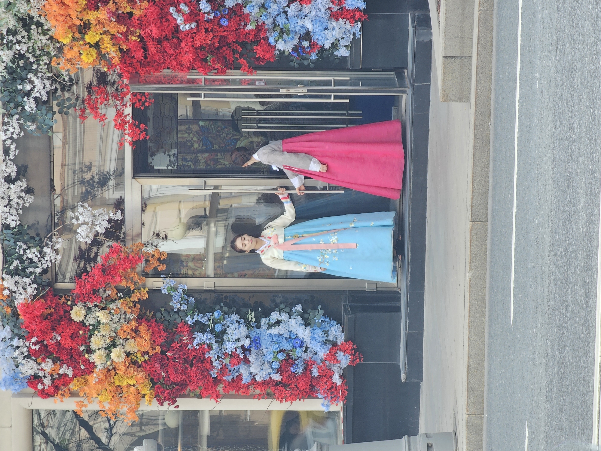 Two women wearing traditional Korean hanboks stand in the open doorway of a building adorned with vibrant flowers. One woman wears a light blue hanbok with a pink sash, and the other is dressed in a pink hanbok with a gray top. The building is located in Moscow, Russia. A sidewalk and street are visible to the right of the building.