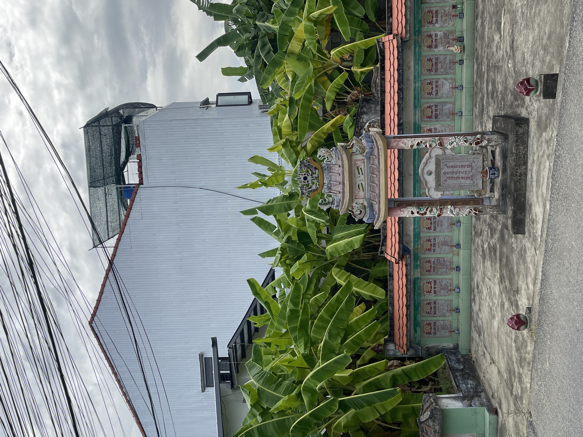 In the city of Hoi An, Vietnam, an outdoor scene features a row of traditional Vietnamese gravestones against a backdrop of lush banana trees and a modern building. The gravestones, set against a wall with an orange tiled roof, are inscribed with names and likely dates, accompanied by small incense burners and offerings. In the foreground, a decorative altar-like structure stands, adorned with intricate carvings and topped with a tiered roof. Two ornamental lotus-shaped objects are placed on pedestals near the altar. A gray asphalt road runs along the bottom of the frame, suggesting this may be near a public thoroughfare or a residential area. The sky is overcast, with a tangle of electrical wires crisscrossing the upper portion of the image, obscuring the view of the sky and the upper parts of the modern building.
