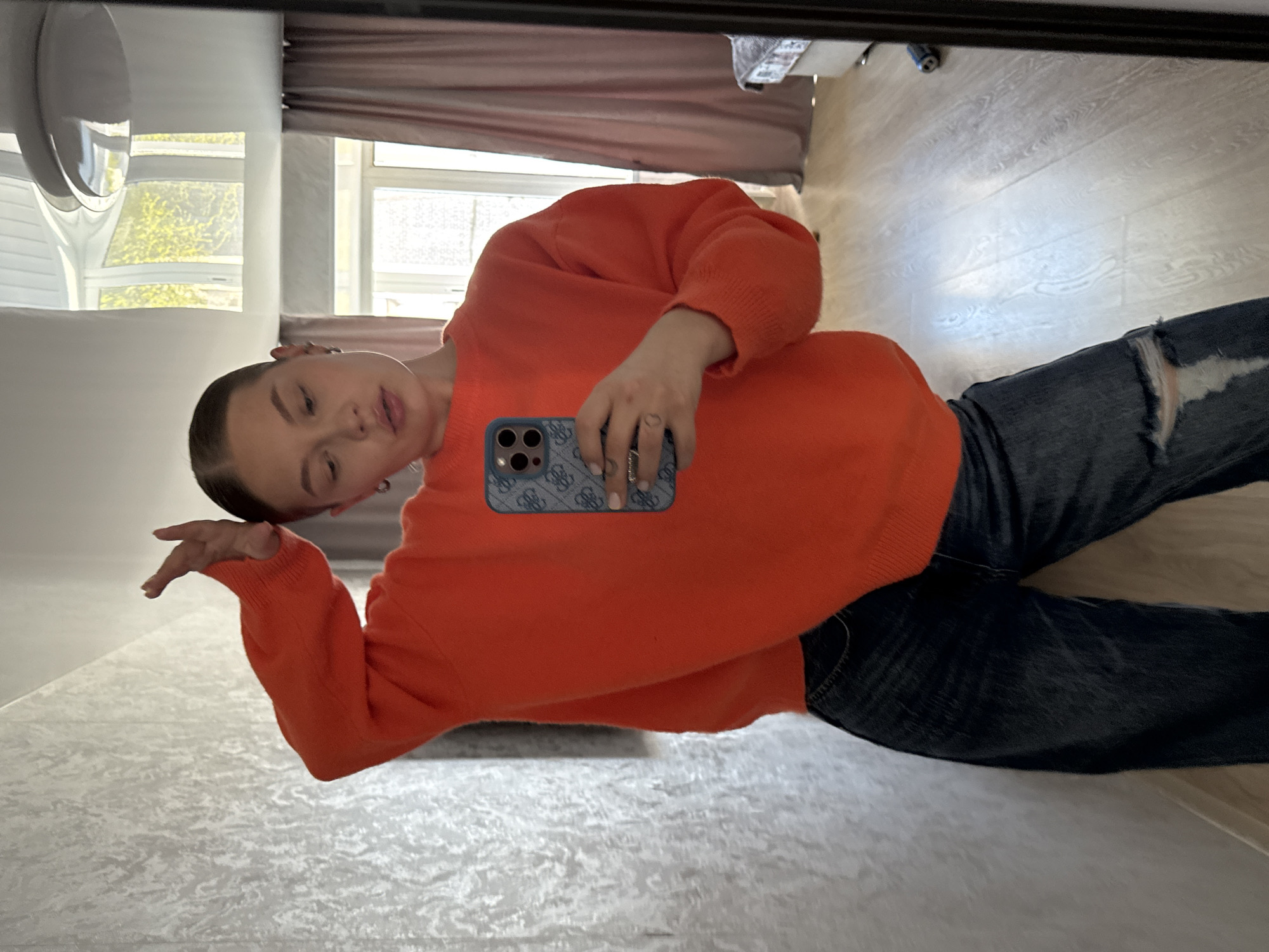 A young woman is taking a selfie in what appears to be an apartment. She is wearing an orange sweater and ripped blue jeans. She is holding a blue cellphone in her left hand. The background includes a window with a view of trees and a building, suggesting daylight. The location is Chaikovsky, Russia, but this detail is not visually discernible from the image itself.