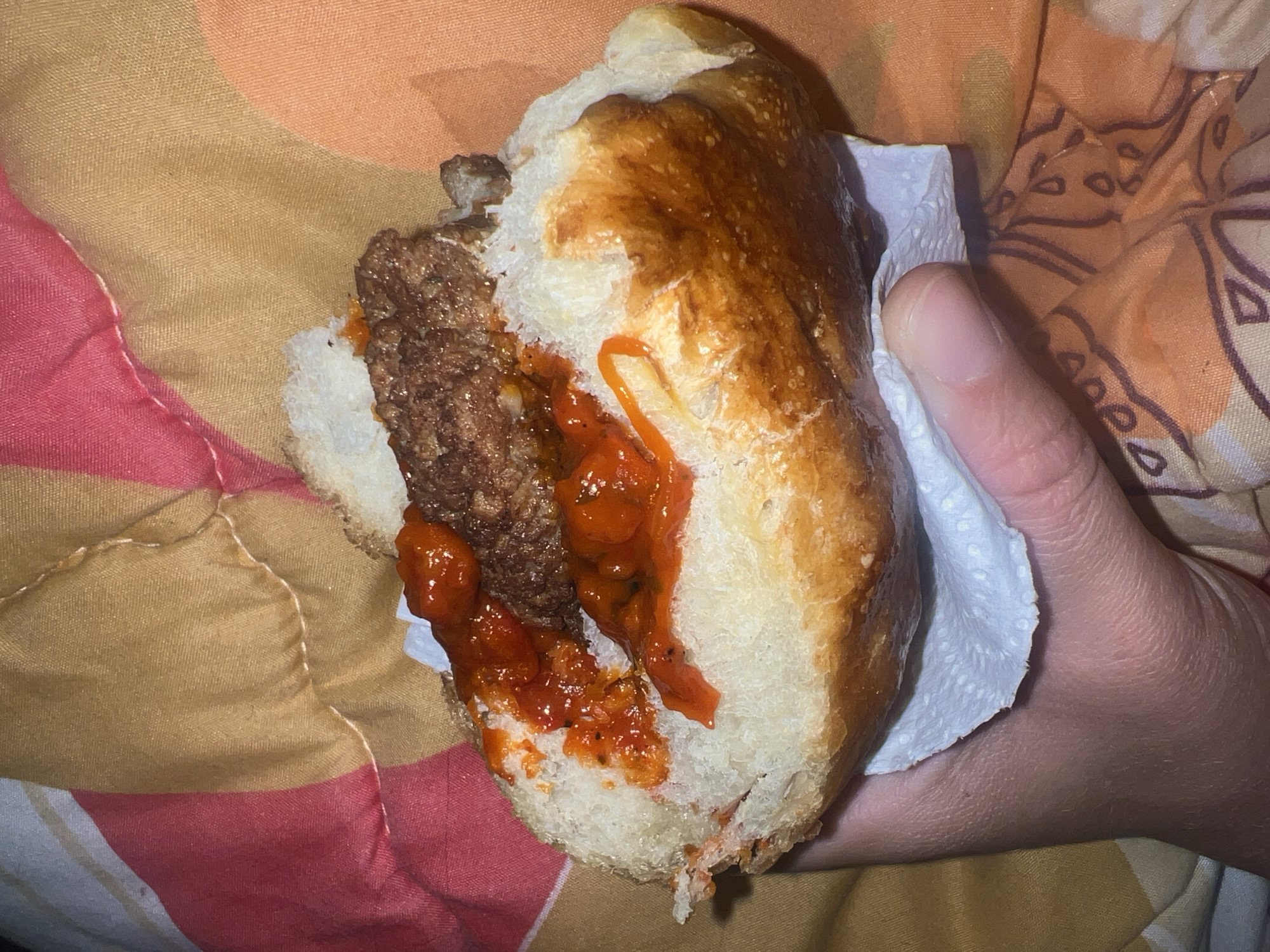 A partially eaten sandwich is held in a human hand against a patterned background. The sandwich consists of a soft, white bread roll filled with a meatball and red sauce. A white napkin is visible between the hand and the sandwich. The background features a pattern of tan, orange, and red shapes on fabric. The location and specific details of the scene are not discernible from the image.