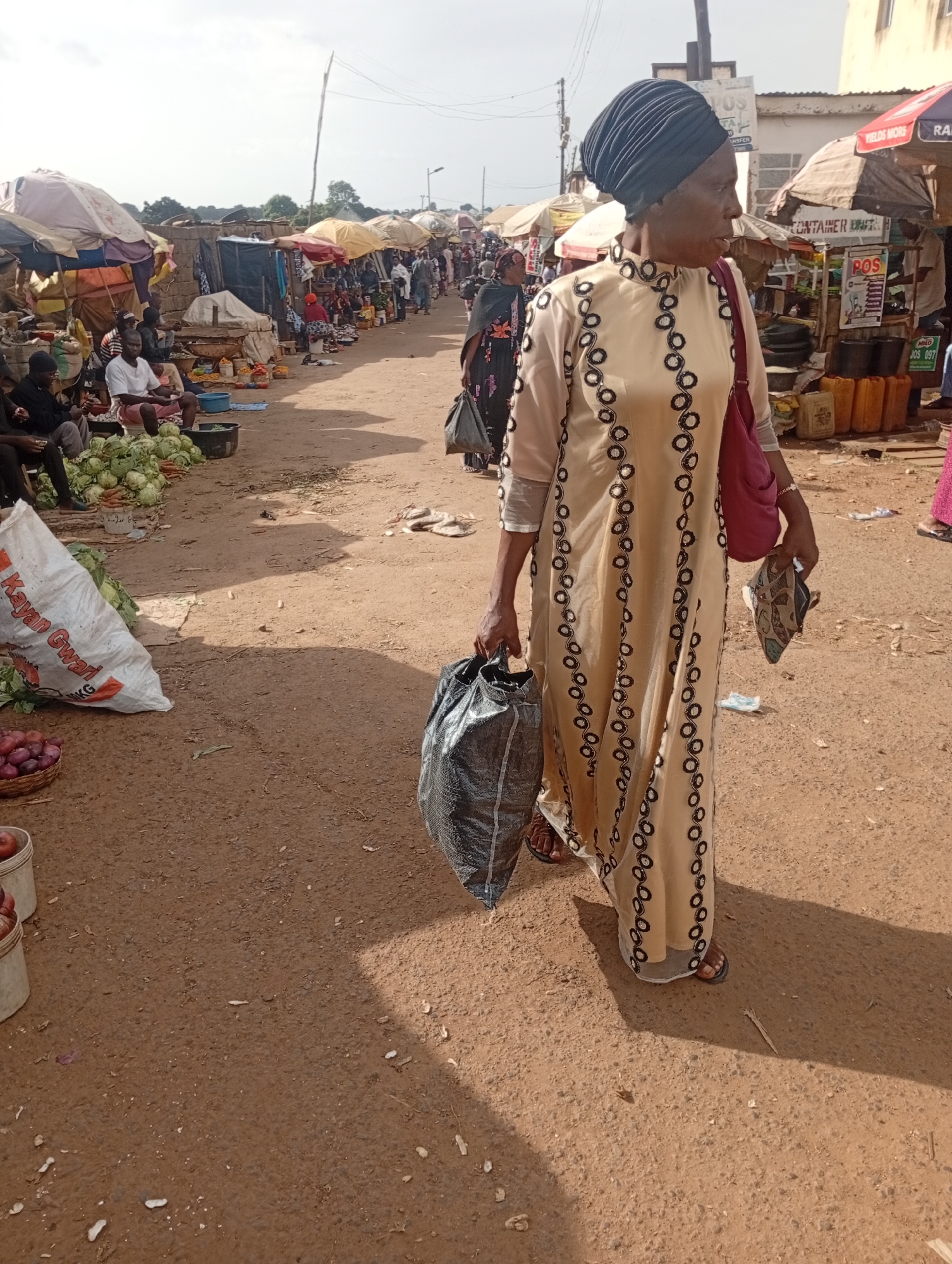 In an outdoor market scene, likely in Jos, Nigeria, a woman wearing a black headscarf and a long, cream-colored dress adorned with circular black embroidery walks forward, carrying two shopping bags and a shoe in her hand. The market is bustling with activity, indicated by the presence of numerous stalls with awnings and various goods displayed, including piles of vegetables like cabbages and onions, and large containers. People are seen sitting and standing around the stalls, suggesting ongoing commerce. The ground is unpaved, dusty, and appears to be a dirt road. The sky is overcast, casting soft shadows. The overall impression is that of a lively and busy local marketplace.