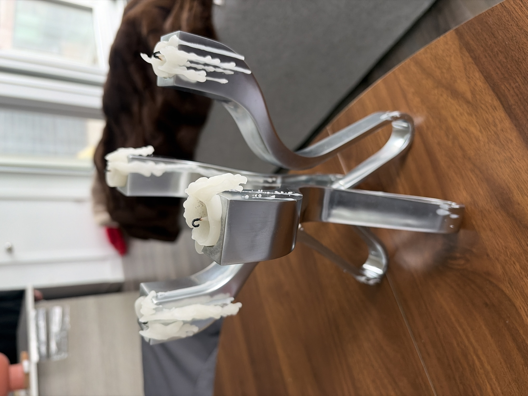 The image depicts a metal candelabra with three partially burned white candles. The candelabra rests on a wooden table with a brown, walnut-like finish. In the background, there is a blurred view of a residential setting, potentially a living room, including a window, a brown furry object possibly a dog or a blanket, a light gray sofa or chair, and a white cabinet. The scene appears to be indoors, likely within a home or apartment. There is no definitive evidence to confirm the location is specifically in Montreal, Canada.