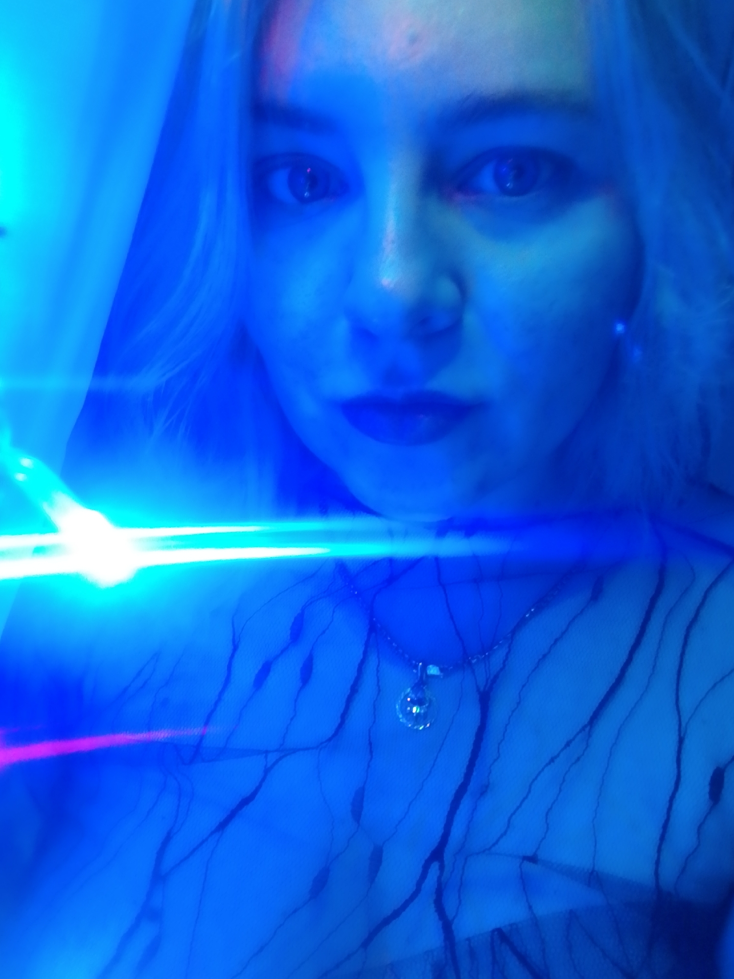 A close-up portrait of a fair-skinned person with light hair illuminated by bright blue light, creating a stark contrast. The person is wearing a dark-colored top with a patterned texture. A silver necklace with a round pendant is visible around their neck. The image includes lens flare, and the background is obscured by light and blur. There is no indication of Izhevsk, Russia.