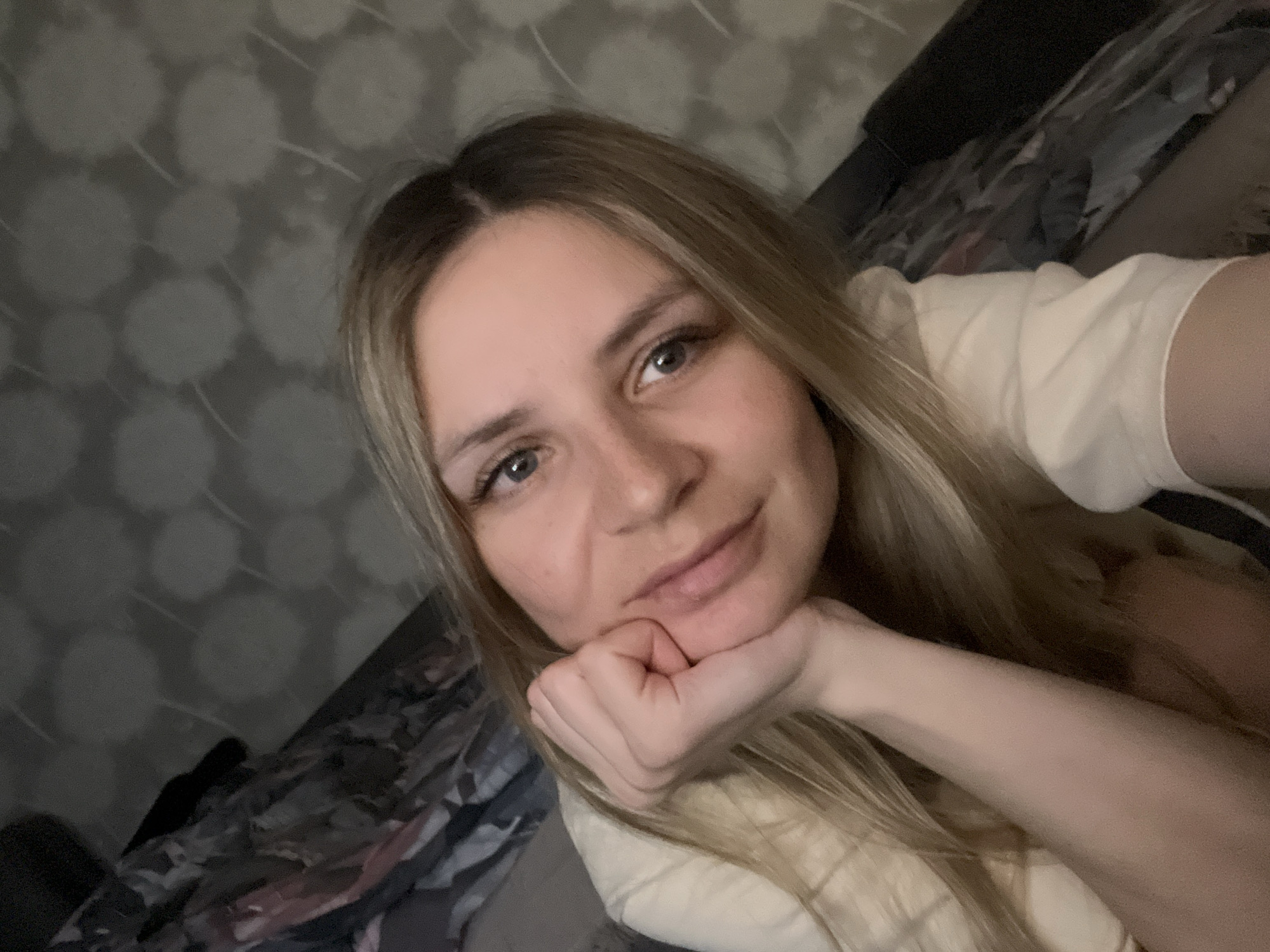 A single individual, a young adult female with long blonde hair and light-colored eyes, is depicted looking directly into the camera. She has a slight smile and is resting her left hand under her chin, with fingers cupped against her cheek. She is wearing a light-colored, long-sleeved garment. The background consists of a wall with dark gray wallpaper featuring a repeating pattern of light, stylized circular elements. To the left and behind her, a section of patterned fabric, possibly bedding or upholstery with dark, abstract designs, is visible. The scene is an indoor, domestic environment. The image does not contain specific visual information to confirm the location as Chaikovsky, Russia.