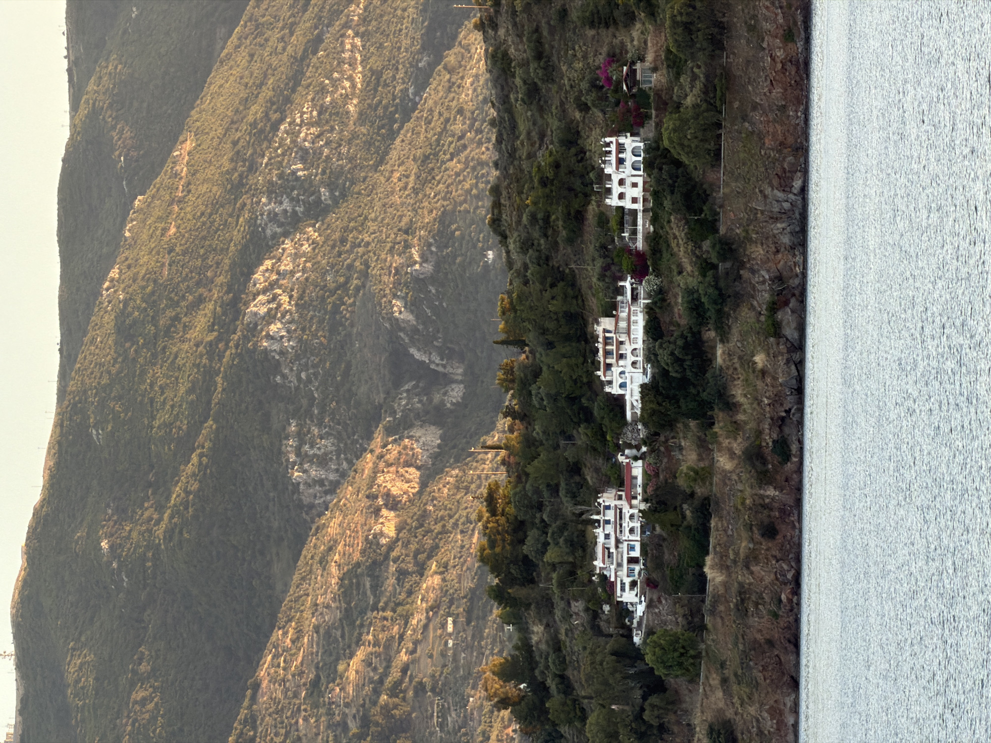 The image displays a serene coastal landscape in Poros, Greece, under the soft light of late afternoon or early evening. The scene is dominated by steep, verdant mountains on the left, their slopes densely covered in green vegetation with visible rock formations catching a warm, golden glow.
Along the base of these mountains, transitioning into the midground, a series of charming white buildings are nestled into the hillside. These multi-story structures, likely villas or houses, exhibit traditional Mediterranean architecture with arched windows, prominent balconies, and terracotta-tiled roofs. Vivid splashes of pink and red flowering plants, possibly bougainvillea, add vibrant color to the green surroundings of the residences. A narrow, winding path or road can be seen connecting some of these properties.
To the right, a calm body of water, presumably the sea or a bay, extends across the frame. Its surface is lightly rippled, reflecting the ambient light with a soft, shimmering effect. The water meets the rugged, rocky coastline at the foot of the hill, which is sparsely covered with shrubs and trees. The overall impression is one of peaceful natural beauty and a picturesque Mediterranean setting. No people or visible text are present in the image.