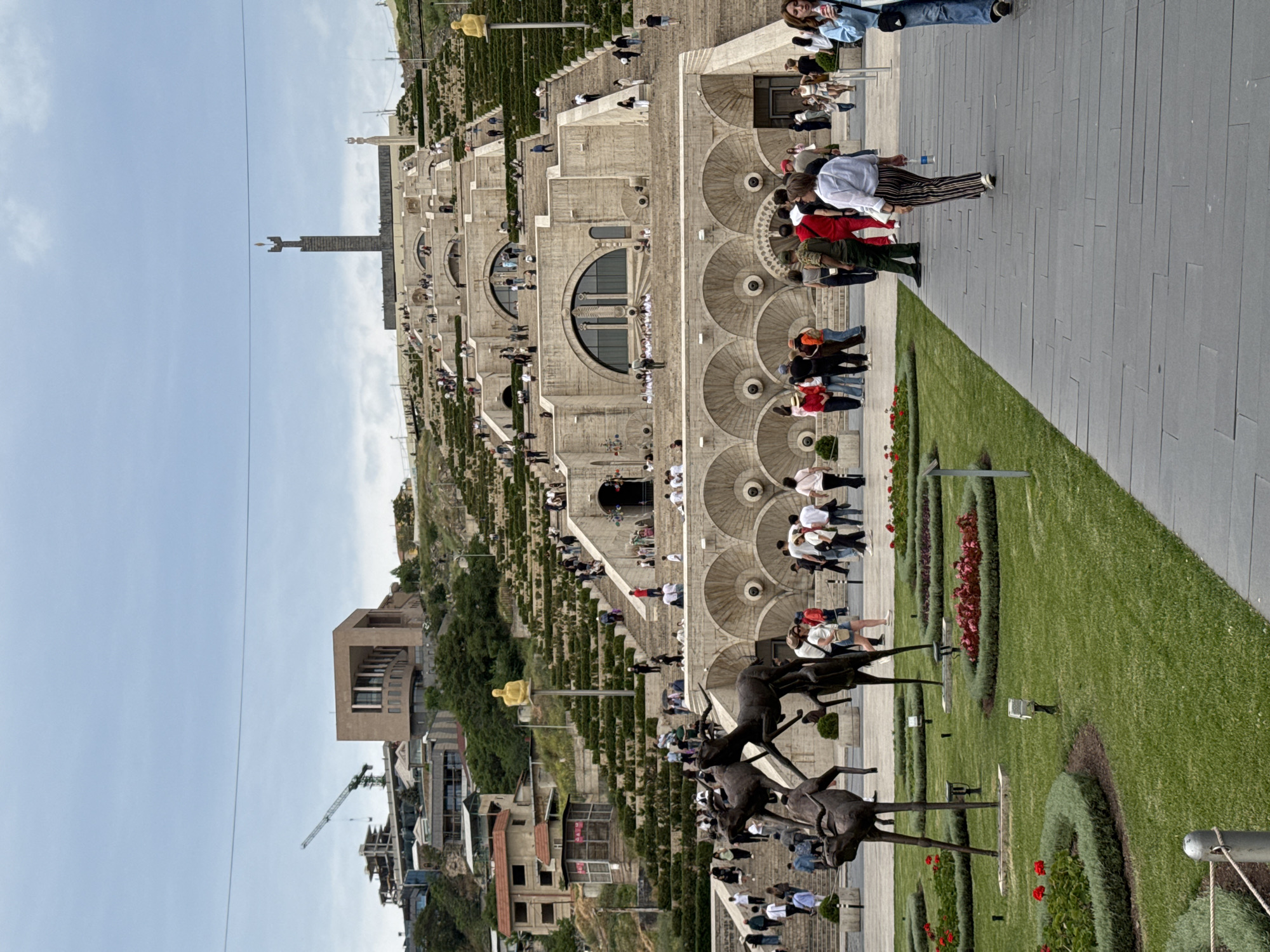 This photo shows the Yerevan Cascade, a large stairway in Yerevan, Armenia. The cascade is a series of tiered levels connected by steps and interspersed with fountains and sculptures. Many people are walking on the cascade and the adjacent plaza. Several sculptures are visible. The buildings on the left side of the photo are typical of Yerevan's architecture. The sky is light blue.