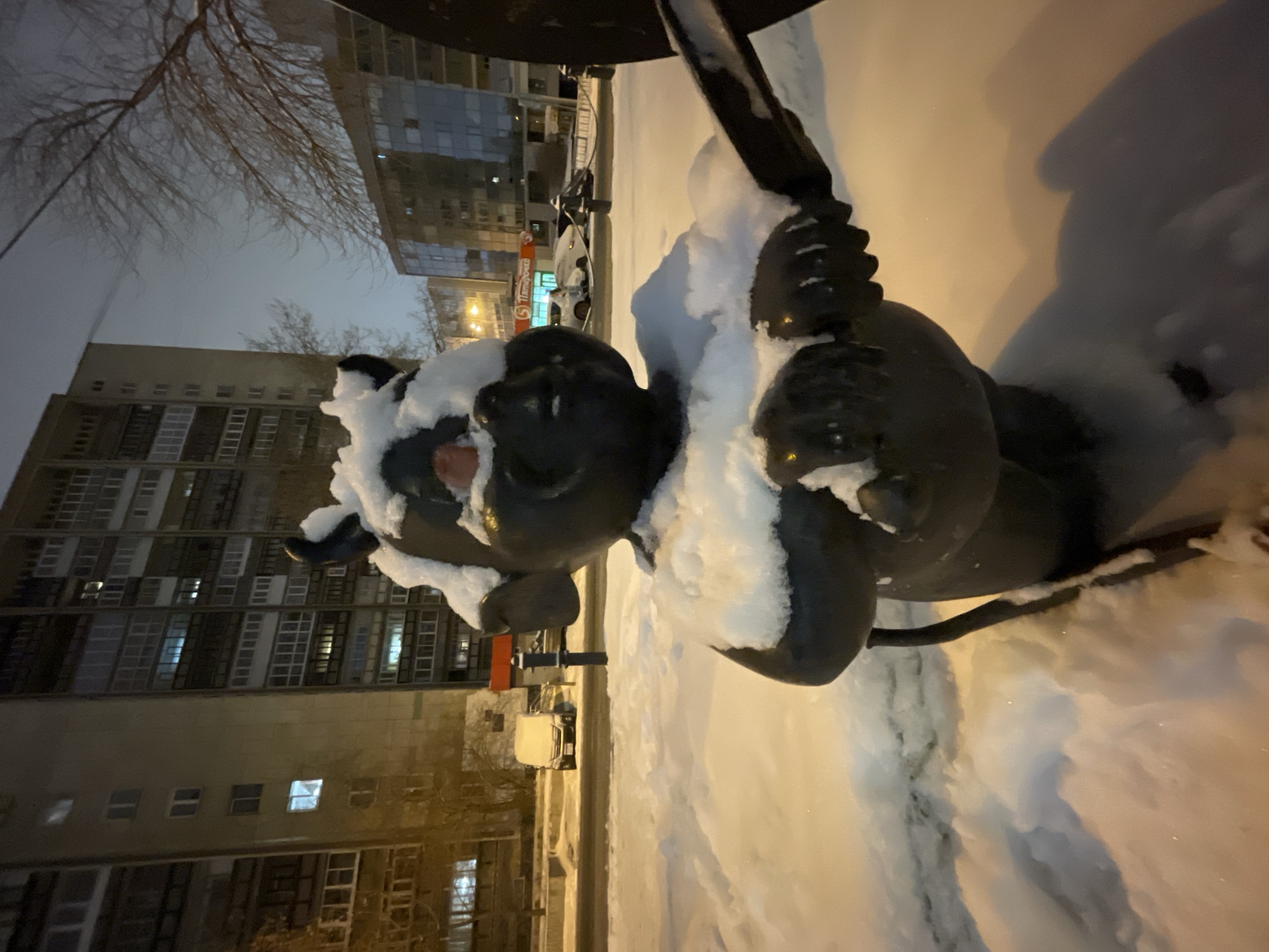 The image shows a bronze statue of a baby angel, partially covered in snow, in Yekaterinburg, Russia. The statue is situated on a snow-covered ground. Buildings are visible in the background. The street is visible with cars parked along it. The lighting suggests it is either dusk or dawn.