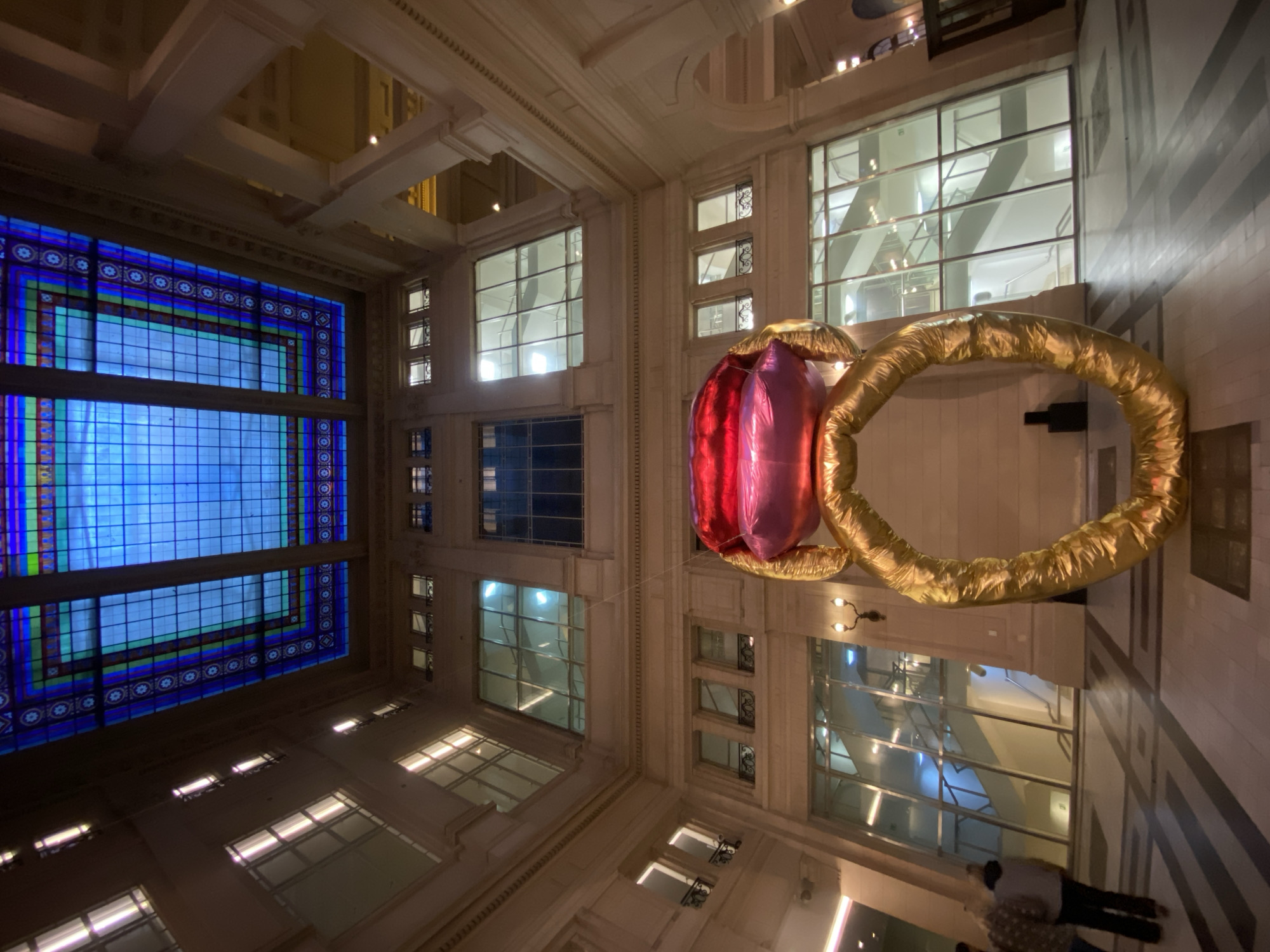 The image depicts the interior of a grand building, possibly a hotel or public space, in Buenos Aires, Argentina, judging by the architectural style. The perspective is from ground level, looking upwards at a distorted angle, showcasing the ornate ceiling and walls. A large, inflatable balloon sculpture in the shape of an engagement ring with a red gem hangs in the center of the frame. The architecture features multi-story windows and detailed molding. Some people stand in the lower right corner, seemingly observing the inflatable ring. The scene is well-lit, highlighting the architectural details and the vibrant colors of the stained glass ceiling.