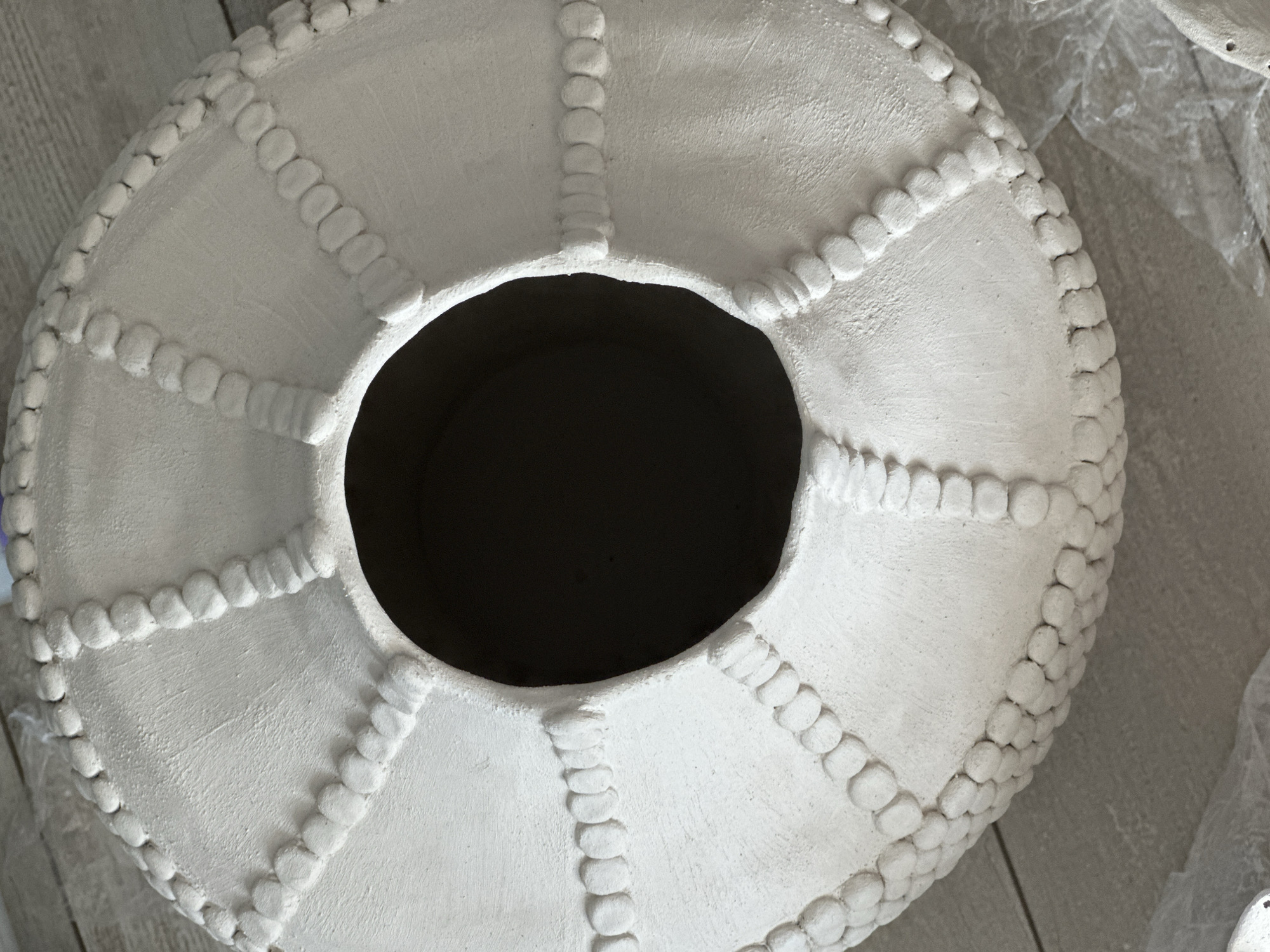 A close-up, overhead shot shows the textured surface of a large, unglazed ceramic vase or pot. The object is white and features a decorative pattern of raised, beaded bands and segmented panels that radiate from a central, dark opening. The texture of the ceramic is rough and matte. The vase sits on a light-colored, possibly wooden floor. The image does not contain any information about the city of Nevinnomyssk, Russia.