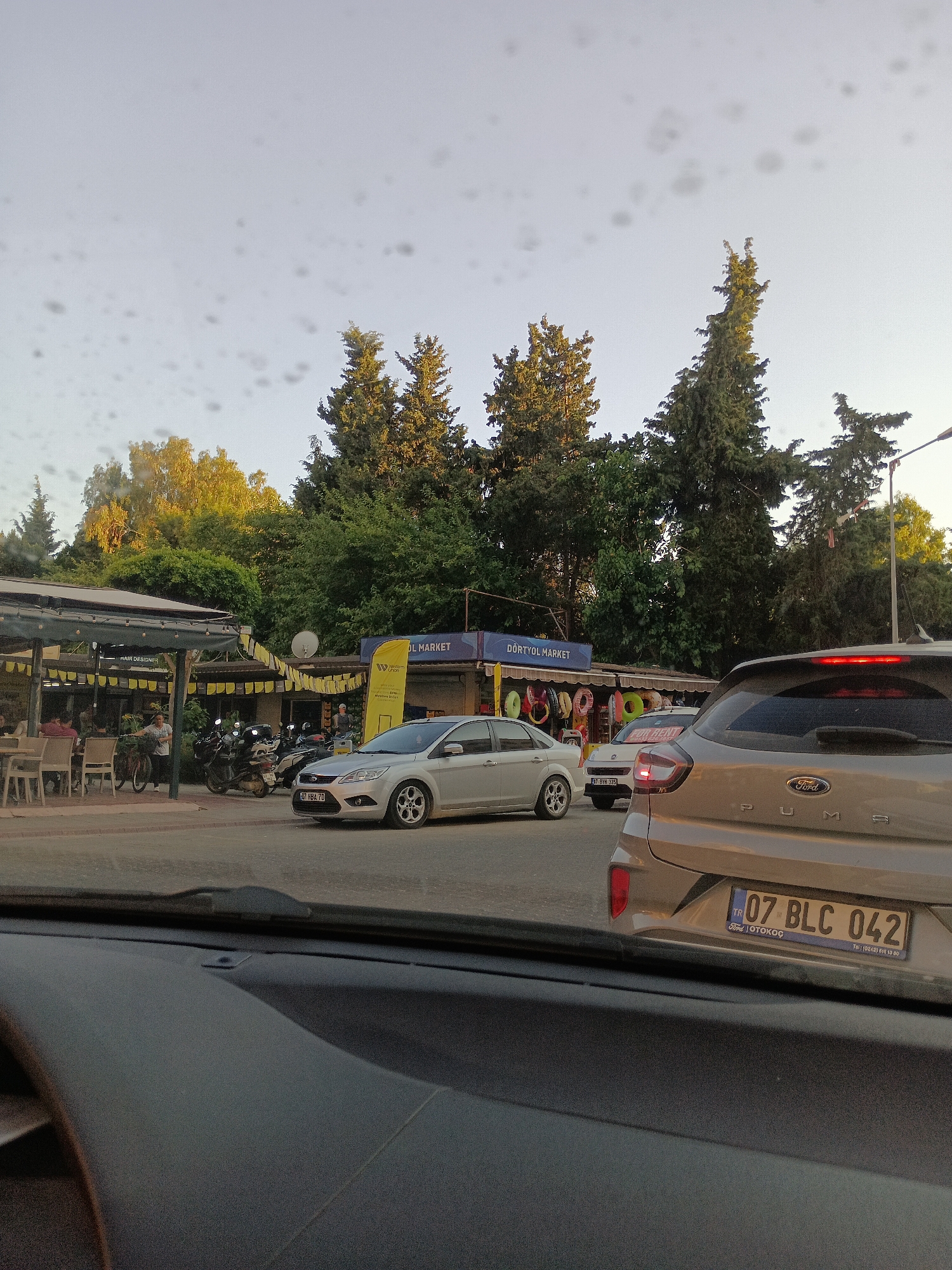 This media file depicts a street scene in Alanya, Turkey, likely during late afternoon or early evening as indicated by the warm, soft lighting and long shadows. The foreground is dominated by the interior of a vehicle, suggesting the photo was taken from a car. The view looks out onto a moderately busy street with several cars stopped, indicating a traffic situation or a pause.
In the middle ground, a silver sedan is parked on the right side of the road, and a beige Ford SUV is in the foreground, viewed from the rear. Behind these vehicles, there's a row of shops and establishments. One prominent sign reads "DÖRTYOL MARKET." Adjacent to it, another stall displays various colorful rings, possibly for sale or as decorations. A covered outdoor seating area for a cafe or restaurant is visible on the left, with a few patrons seated and a person standing near a bicycle. Several motorcycles are parked in front of the market.
The background is filled with lush green trees, with some showing hints of yellow from the setting sun. The sky is a pale, clear blue, with some circular spots that might be water droplets or imperfections on the camera lens. The overall atmosphere appears calm and everyday, typical of a street in a Turkish city. The visible text includes shop names and license plate numbers (e.g., "07 BLC 042," "07 HBA 70," "07 BYH 335"), which can help identify the specific location. The presence of the Ford dealership sticker on the SUV's license plate frame further confirms its origin and potential context.