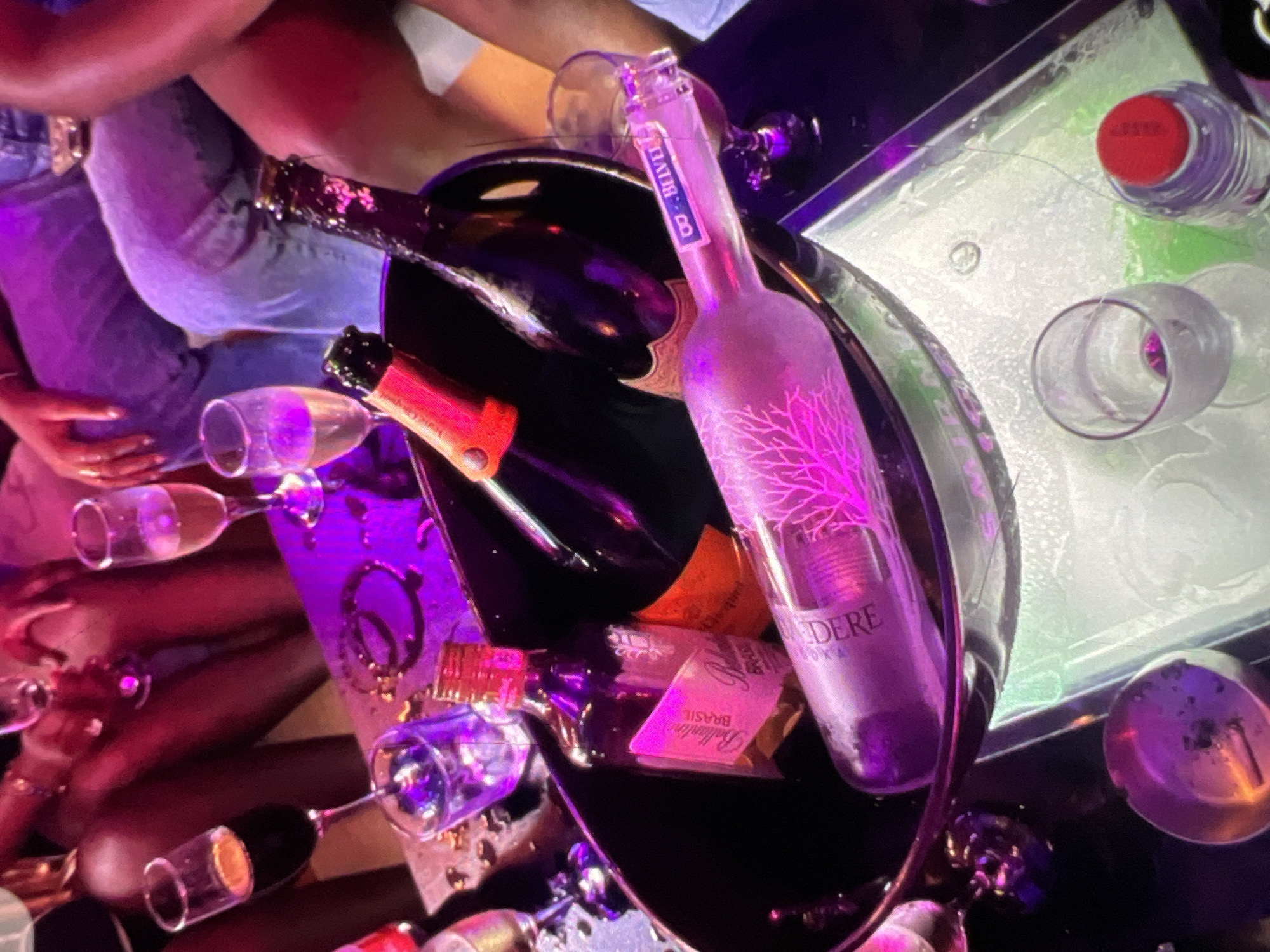 This image depicts a scene at a nightclub or lounge in Lagos, Nigeria. The focus is on a black ice bucket filled with several bottles of alcohol, including what appears to be champagne and vodka. The bottles are illuminated by a vibrant purple and pink light, creating a lively atmosphere.
Several individuals are present, though their faces are not clearly visible. They are seen in the background, with parts of their bodies, including arms and legs, visible. Some are holding glasses, suggesting they are engaged in drinking and socializing. The presence of champagne flutes and other glassware indicates a celebration or a night out.
The environment appears to be dimly lit, typical of a nightclub setting, with colored lights contributing to the mood. A table with a reflective surface is visible in the foreground, holding the ice bucket and some empty glasses. An ashtray is also present, suggesting smoking may be occurring in the venue. The overall impression is one of a social gathering in a nightlife establishment.