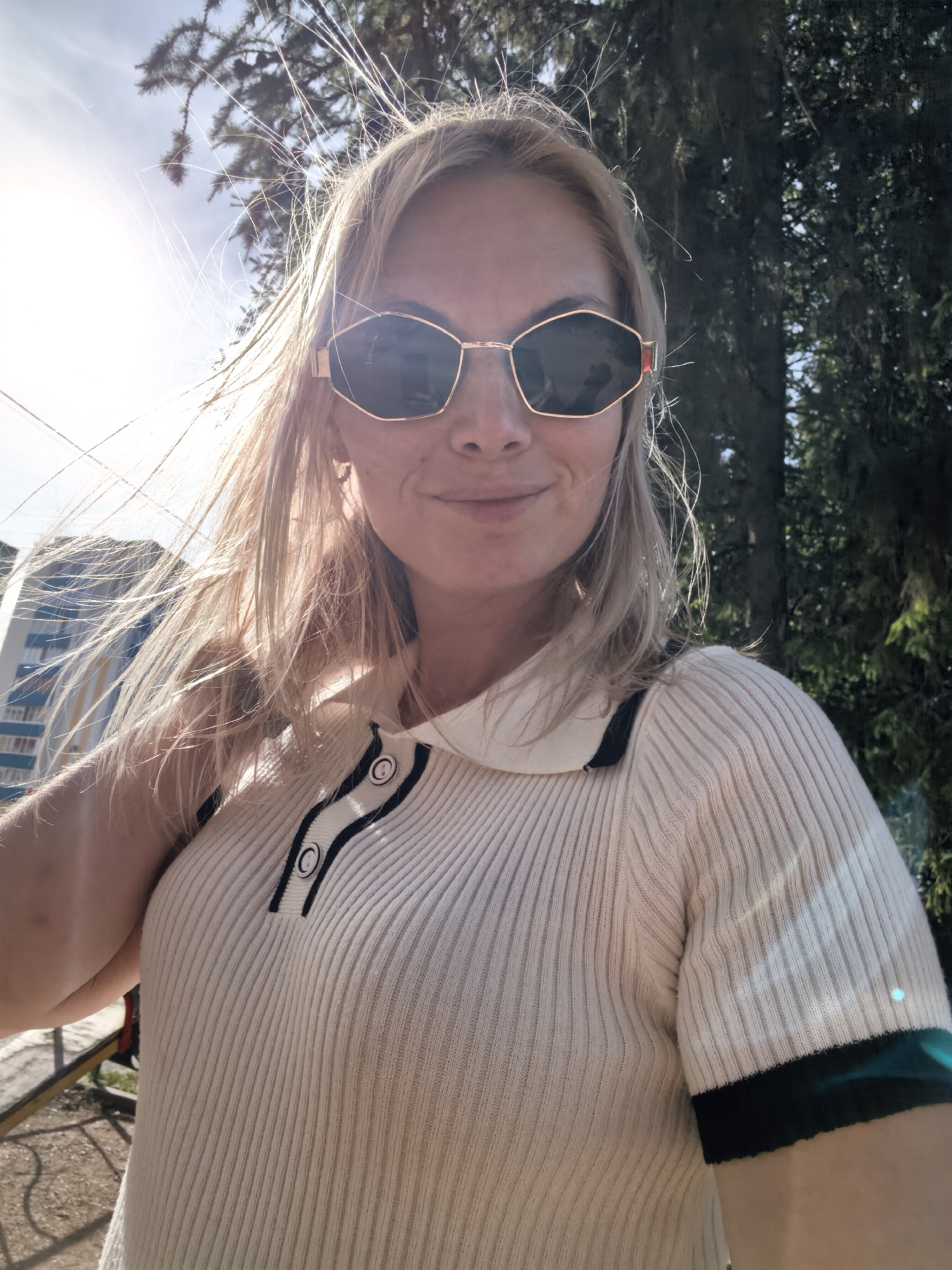 A blonde woman with sunglasses is taking a selfie outdoors in Sterlitamak, Russia. She is wearing a cream-colored ribbed shirt with black and white trim on the collar and sleeves. Her hair is blowing in the wind. In the background, there are trees and a building. The sun is bright and casting a glare.