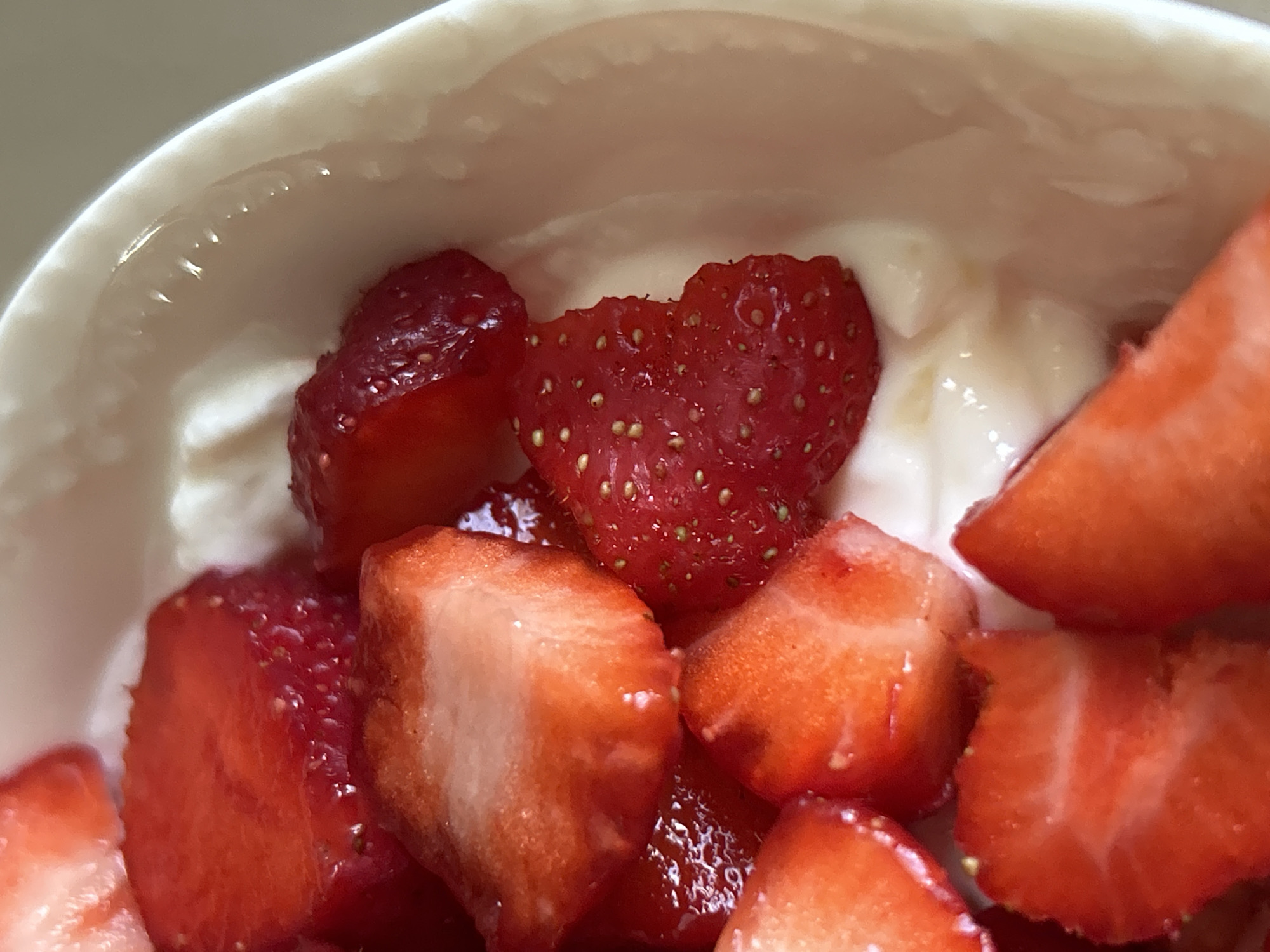 The image is a close-up, overhead shot of sliced strawberries in a white bowl filled with a creamy white substance, likely yogurt. The strawberries are vibrant red, some cut into wedges and others into smaller pieces, revealing their juicy interiors. The bowl's curved rim is visible on the right side of the frame, showing a subtle texture. The lighting is bright, highlighting the textures and colors of the fruit and the creamy base. This image does not provide any information to suggest a location in Plovdiv, Bulgaria.