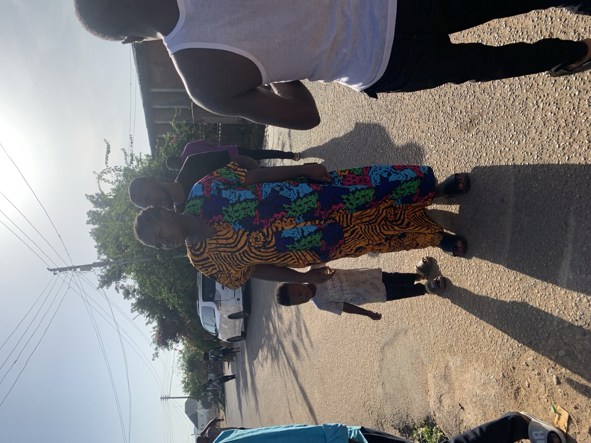 The photo shows a group of people standing on a paved road in Gura Top, Nigeria. A woman in a colorful patterned dress holds the hand of a child wearing a t-shirt and pants. A man in a white tank top and black pants stands nearby. Power lines are visible overhead, and a white van is parked on the side of the road. Buildings are visible in the background. The shadows suggest it is daytime.