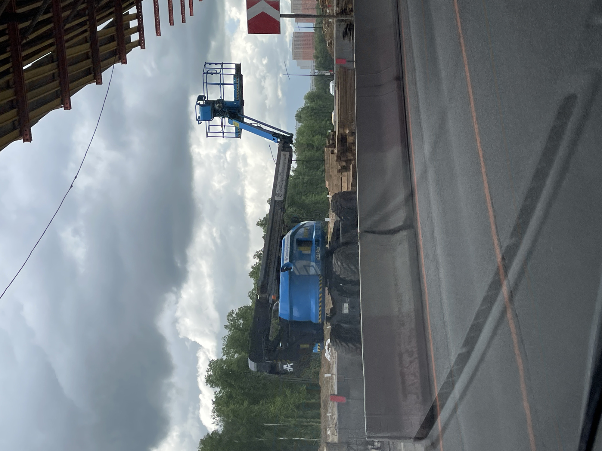 The image shows a Genie S-65 boom lift at a construction site in Novodrozhzhino, Russia. The lift is blue and positioned next to a road. A red and white road sign is visible in the background, along with construction materials and trees. The sky is overcast with dark clouds. A building structure is partially visible on the left side of the image. The photo appears to have been taken from inside a vehicle, with a window frame partially visible on the right.
