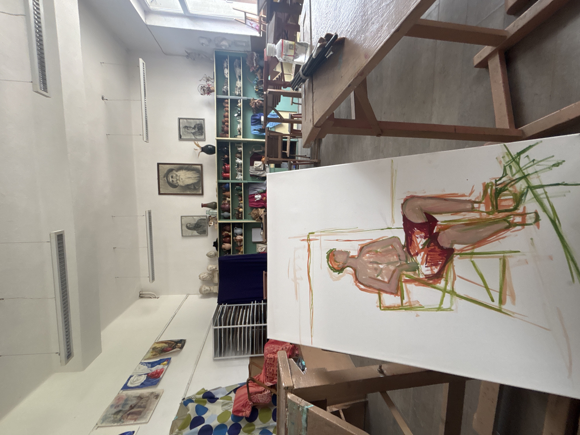 The image shows an art studio in Troyan, Bulgaria. A large canvas dominates the foreground, displaying a sketch of a reclining figure in red and green hues. The canvas is set on an easel, which is partly visible at the bottom of the frame. Behind the canvas, a wooden table is visible, holding a variety of art supplies. The studio itself appears spacious, with a high ceiling and natural light. A tall shelf lines the back wall, filled with diverse objects and artworks. Paintings are arranged on the adjacent wall, and a rack filled with artwork sits near a draped surface. The overall impression is of a working art space.