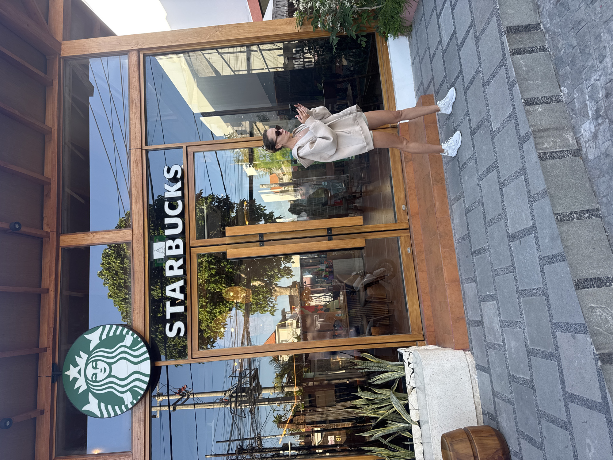 A medium-shot, full-body outdoor image presents a woman in front of a Starbucks in North Kuta, Indonesia. The storefront features vertical wooden doors. The Starbucks logo, a green circle with a stylized mermaid, is affixed to the left of the entrance. The brand name, "STARBUCKS," is vertically printed in white on the glass. The woman, wearing sunglasses and a beige outfit with white sneakers, stands at the entrance. Paved stone flooring is visible in the foreground. Electrical wires stretch across the upper left portion of the image. The background shows reflections of buildings and foliage on the glass. Plants adorn the base of the building on the left and right sides of the entrance. A stone border separates the Starbucks entrance from the sidewalk.