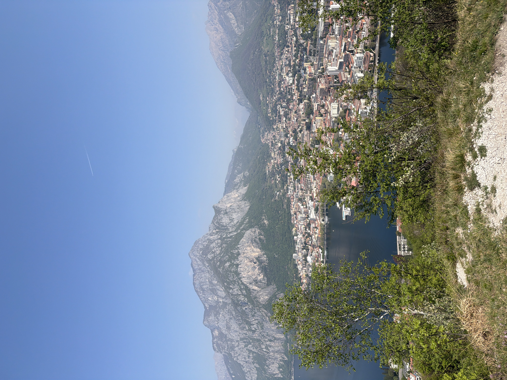 The image shows a hillside view of the town of Galbiate, Italy. The town is nestled between a lake and a large mountain range. The blue sky is clear except for the white trail of an airplane. A grassy hillside is in the foreground with foliage.