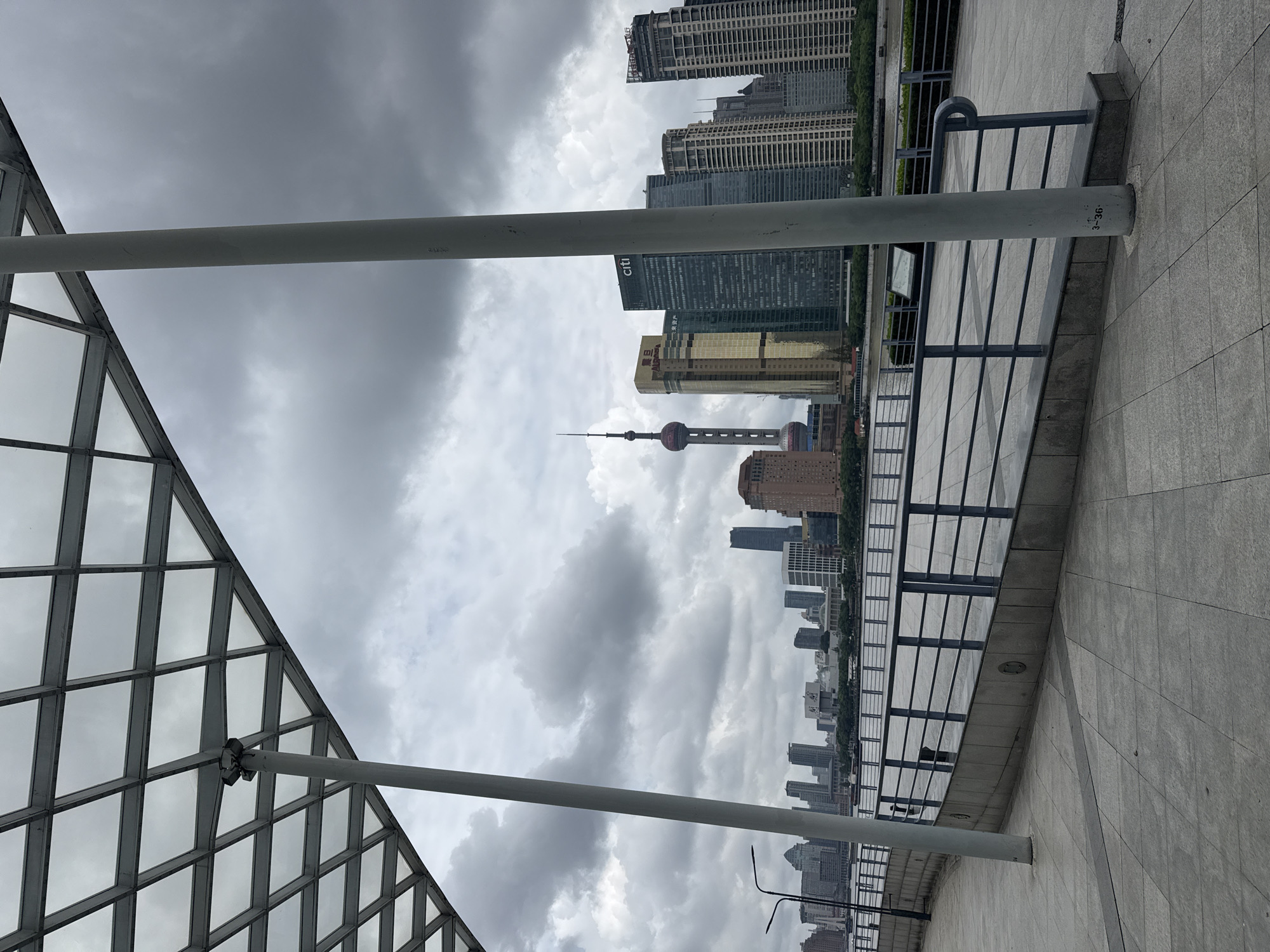 The image depicts a view of the Shanghai skyline from the Bund waterfront promenade on a cloudy day. In the foreground, a portion of a modern, cantilevered roof structure with a metal grid ceiling is visible, supported by thick white pillars. The promenade itself is paved with large, grey square tiles, and a dark grey metal railing runs along the edge, separating the walkway from a body of water.
Beyond the railing, the Shanghai skyline is dominated by numerous skyscrapers. The most prominent structure is the Oriental Pearl TV Tower, a distinctive spherical tower with spires. To its right are several modern high-rise buildings, including the golden Jin Mao Tower and the green Citi building. Further in the distance, more buildings of varying heights and architectural styles form a dense urban landscape. The water in the foreground is calm, reflecting the overcast sky and the surrounding buildings. The overall atmosphere is one of urban grandeur under a subdued, cloudy sky.
