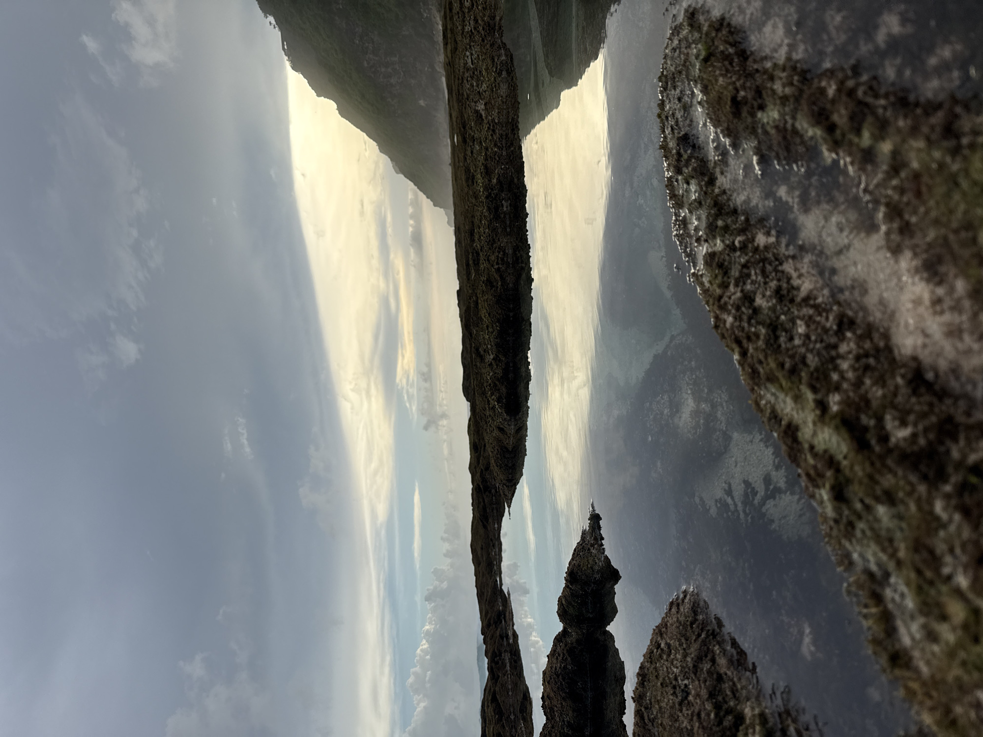 The image shows Uluwatu, Pecatu, Indonesia. The camera faces a cliff edge with rock formations covered in vegetation in the foreground. The sky is visible with clouds, partially illuminated by the sun. A sliver of green mountain can be seen in the background. The ocean is visible between the foreground rocks and the mountain.