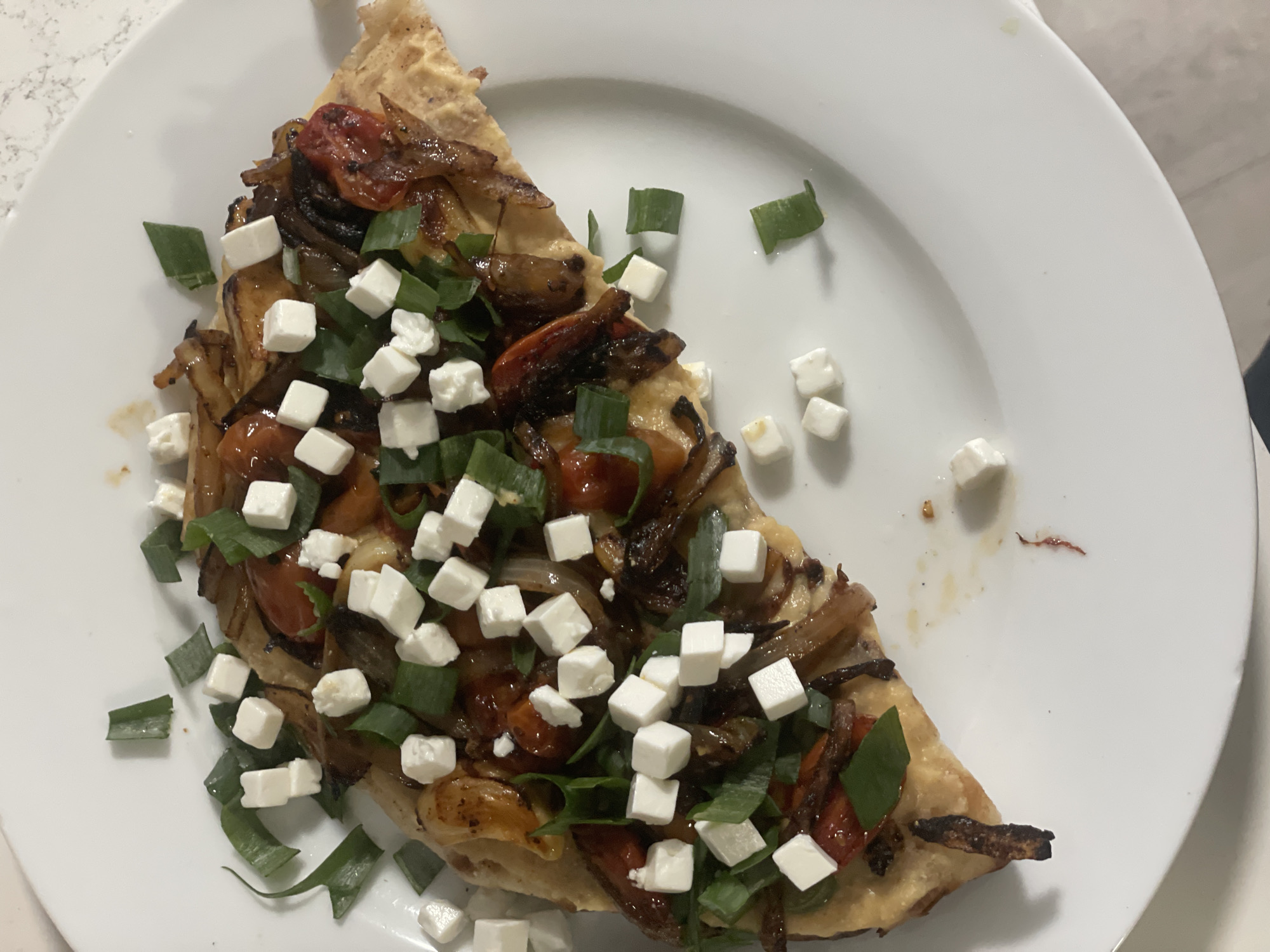 This image presents a close-up view of a single slice of savory tart or flatbread, artfully arranged on a clean, round white ceramic plate. The food item is generously adorned with an array of toppings. The base, appearing as a light brown, possibly thin crust, is covered with glistening, golden-brown caramelized onions, providing a rich, sweet foundation. Interspersed among the onions are soft, reddish pieces that resemble roasted cherry tomatoes, adding a pop of color and a hint of acidity.
Scattered liberally across these cooked vegetables are numerous small, uniformly diced cubes of white cheese, likely feta, which stand out against the darker toppings. Completing the presentation are bright green, freshly chopped pieces of green onions or chives, sprinkled over the entire slice, lending a vibrant color and fresh aroma. A few cubes of cheese and green onion pieces have fallen or been intentionally placed on the white plate around the main food item, enhancing the visual appeal.
The background of the shot shows a light-colored, speckled countertop, suggesting an indoor setting, likely a kitchen or dining area. No people are visible, and there is no discernible text in the image. The scene is brightly lit by ambient indoor lighting. This culinary creation is presented in Raanana, Israel.