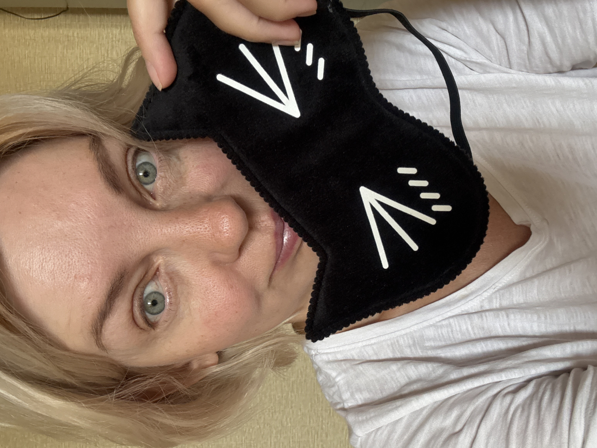 A close-up shot captures a fair-skinned woman with short blonde hair and blue eyes. She is wearing a white t-shirt and holding a black cat-shaped sleep mask to the right side of her face. The mask features white embroidered cat ears and whiskers. The woman is looking directly at the camera, and her mouth is slightly parted. The background appears to be a textured wall. The location is not specified as Sochi, Russia.