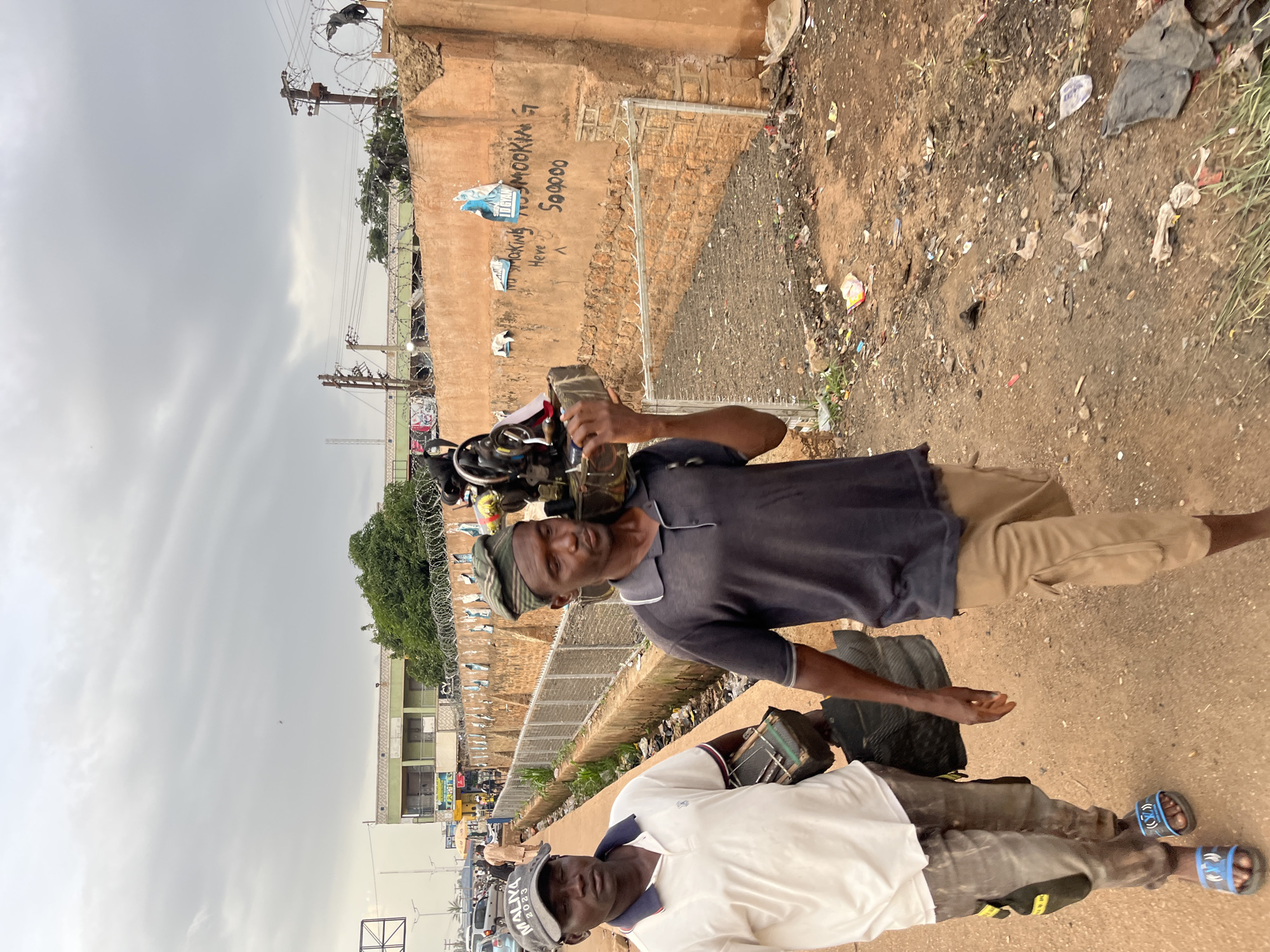 Two men are walking side-by-side on a dirt path in Bukuru, Nigeria, under an overcast sky. The man on the left, wearing a white polo shirt with navy and red trim, a grey baseball cap that reads "MALIYA 2023", and light brown cargo pants with yellow accents, carries a small, dark green box with metal clasps in his right hand. He is also wearing blue and black slides. The man on the right, wearing a dark grey polo shirt and tan cargo pants, carries a sewing machine on his right shoulder. The machine is mounted on a wooden base and appears to have several components attached. He wears a green patterned bandana on his head. Behind the men, a low concrete wall stretches into the background, topped with barbed wire. The wall is covered in various graffiti tags and posters. The ground is littered with trash, including plastic bags, wrappers, and debris. In the distance, buildings and a street with cars and pedestrians are visible.