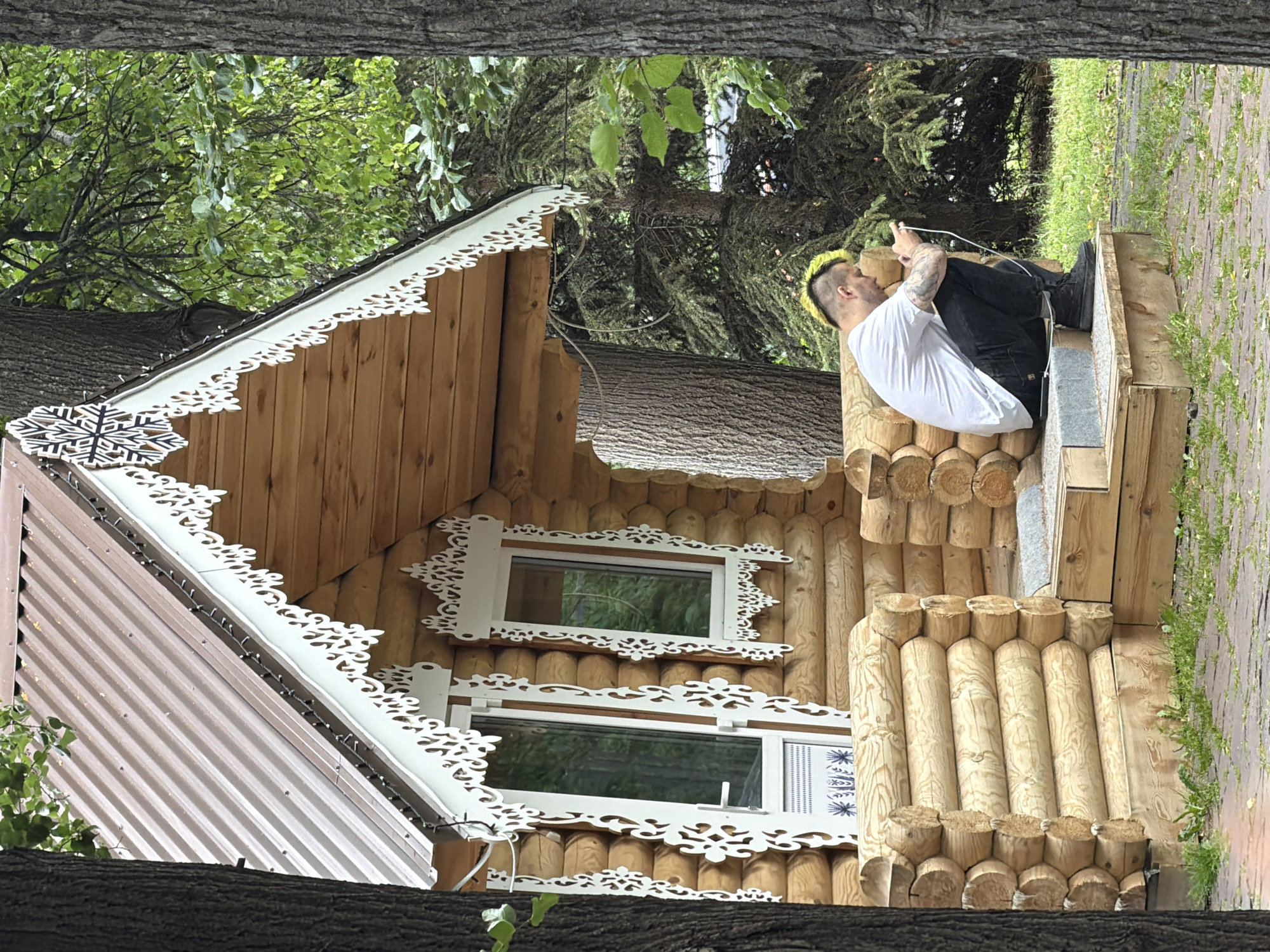 The image depicts a man with bright yellow-green hair, wearing a white t-shirt and black pants, sitting on a wooden structure adjacent to a rustic wooden house. The house features decorative white trim around its windows and along the roofline, resembling snowflakes and intricate floral patterns. The roof is partially covered with brown corrugated metal. The man is looking down at a white object, possibly a phone, in his hands. He has tattoos on his arms. The scene appears to be outdoors, with trees and greenery in the background, suggesting a natural setting. The ground is paved with bricks and there is grass growing between them. The lighting suggests it is daytime, with soft, diffused light indicating an overcast or shady condition. The overall impression is one of a peaceful, possibly rural, environment. The image is oriented vertically.